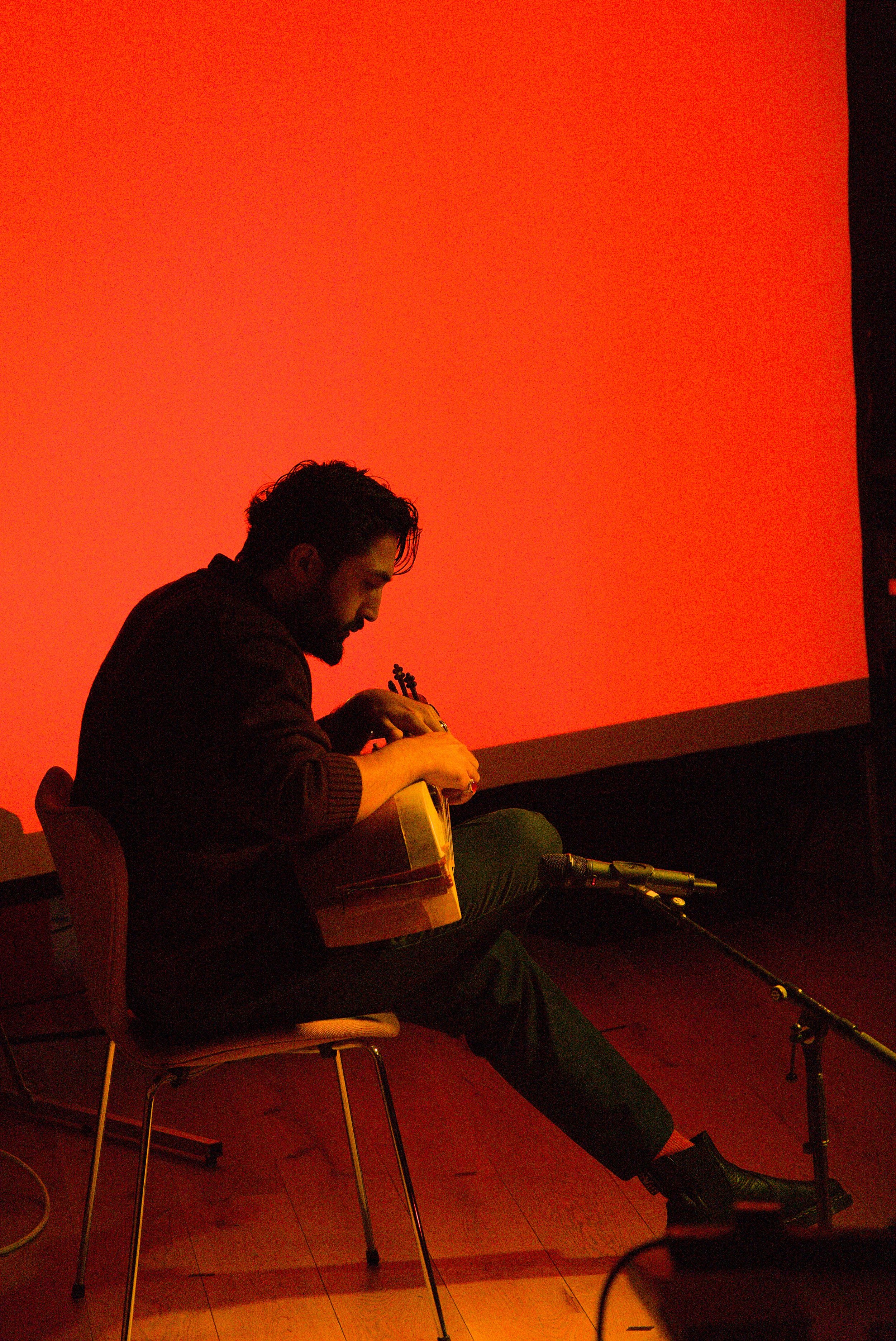A man with dark hair and a beard sitting on a wooden chair, playing an acoustic guitar, with a microphone in front of him, against an orange wall.