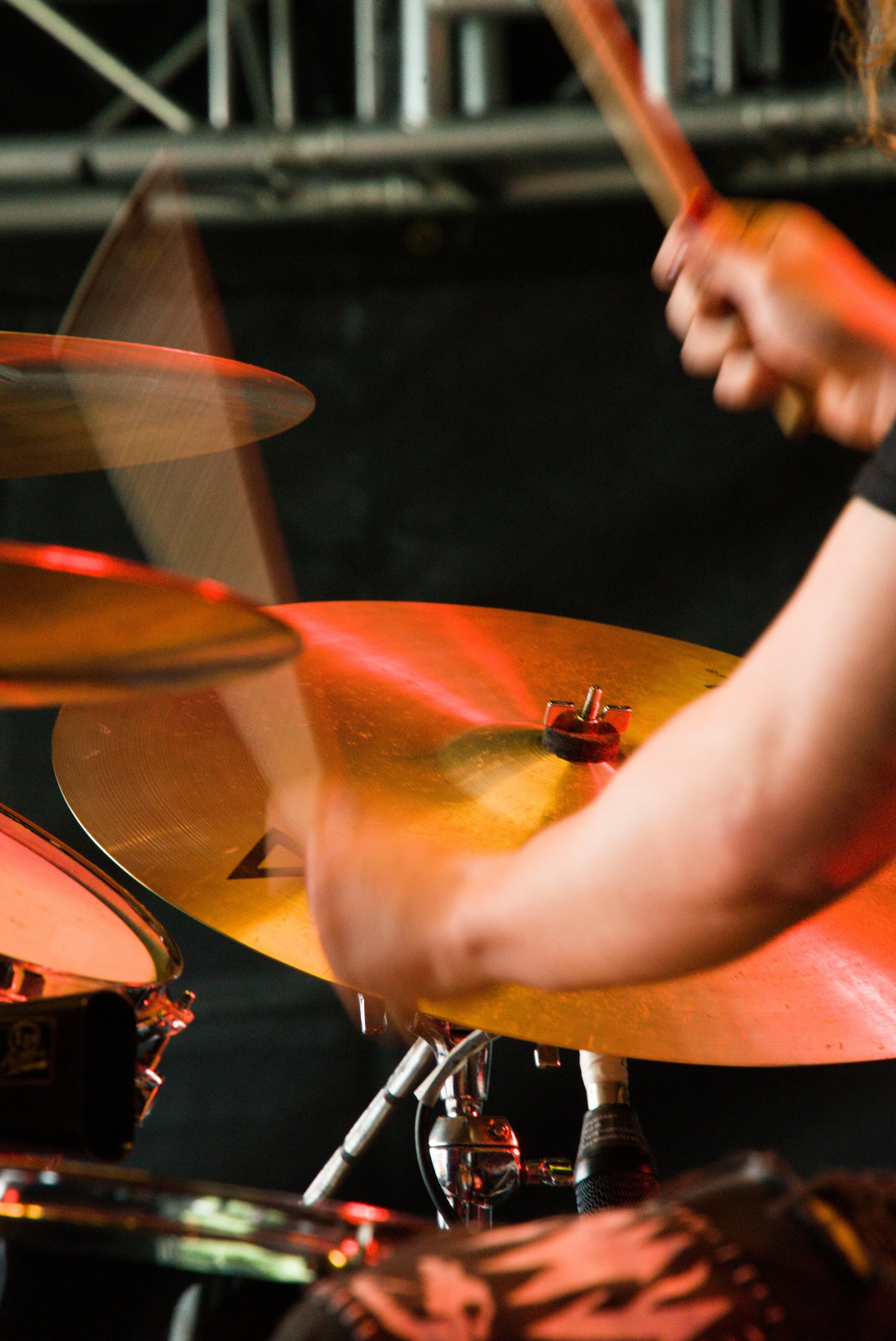 Close-up of a person's arm playing a drum set, with cymbals and drumsticks visible, in a dimly lit setting.