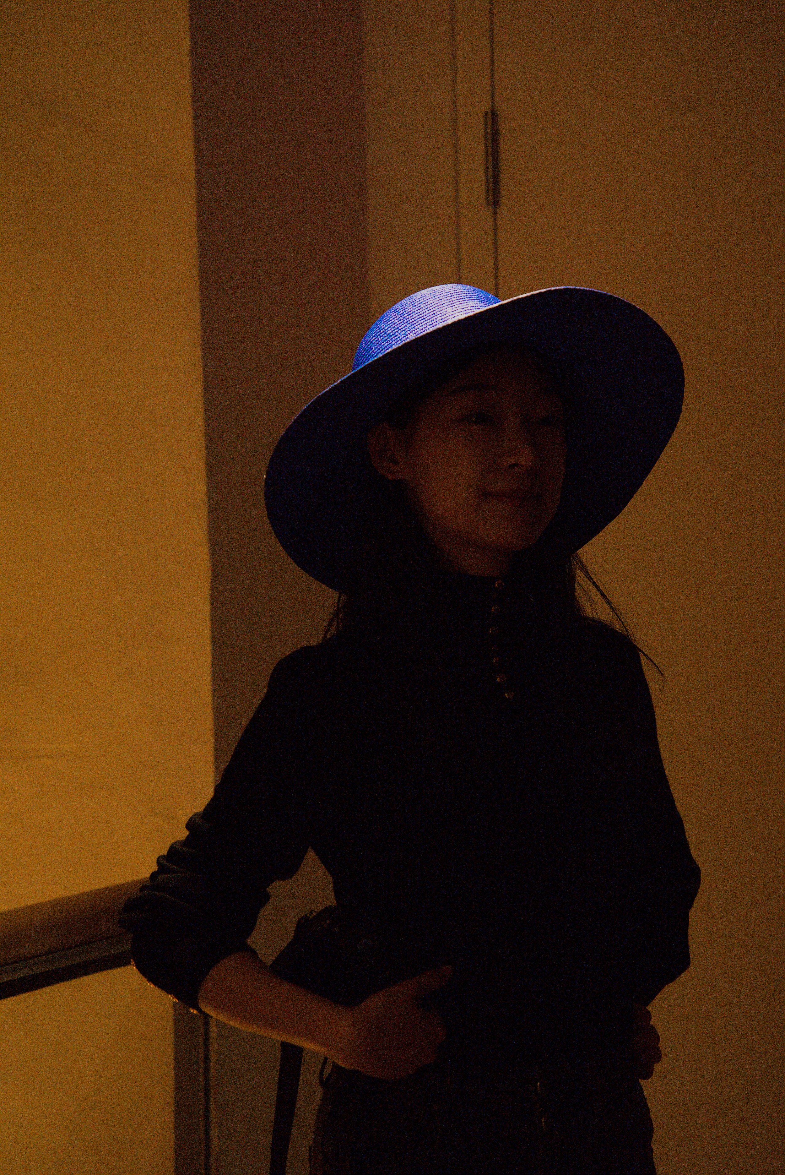 A woman wearing a large wide-brimmed blue hat and dark clothing, smiling softly, standing indoors near a dark corner with a yellow wall.