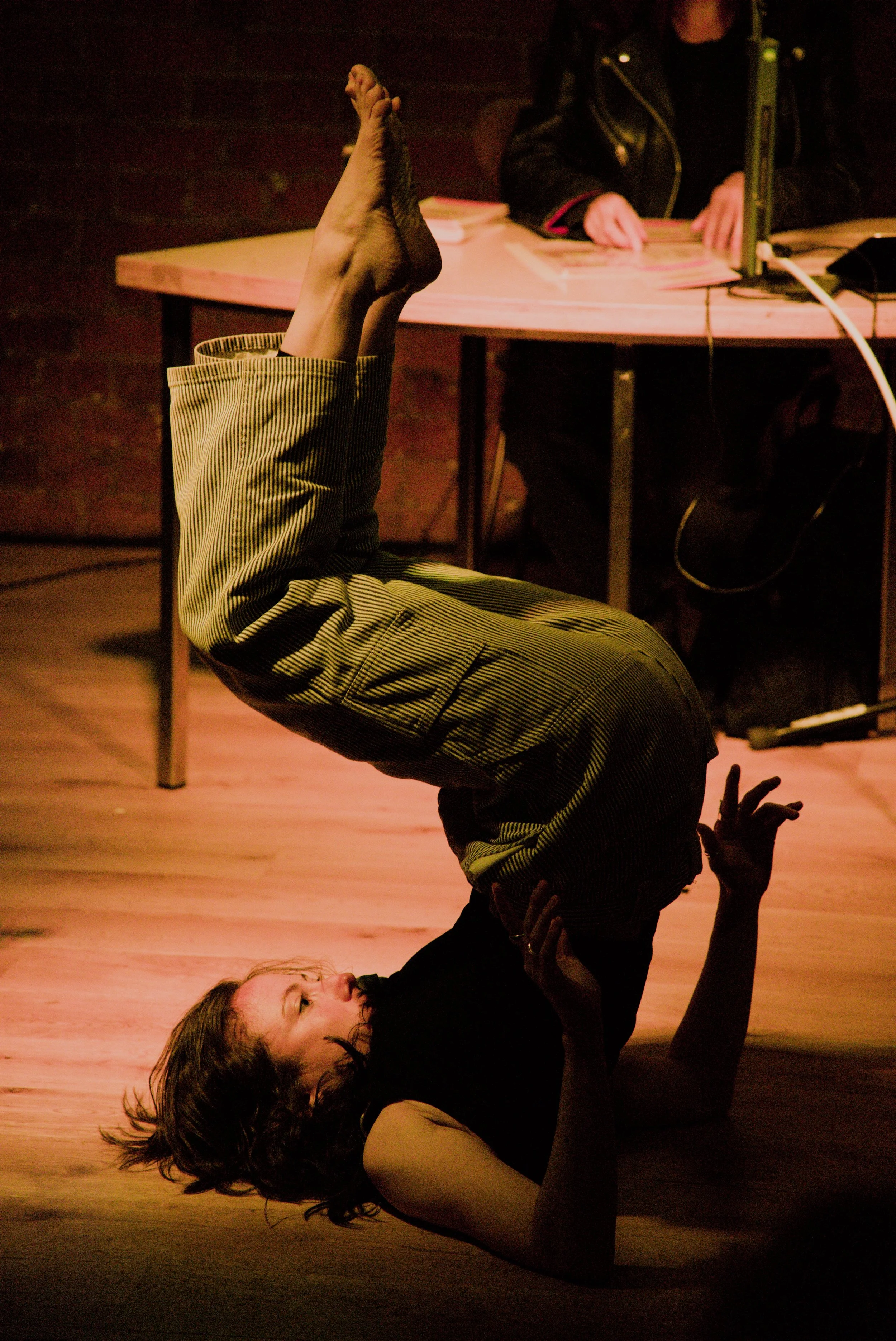 A woman performing an acro yoga pose with a man lying on the ground, supporting her by holding her legs in the air, in a dimly lit indoor setting.