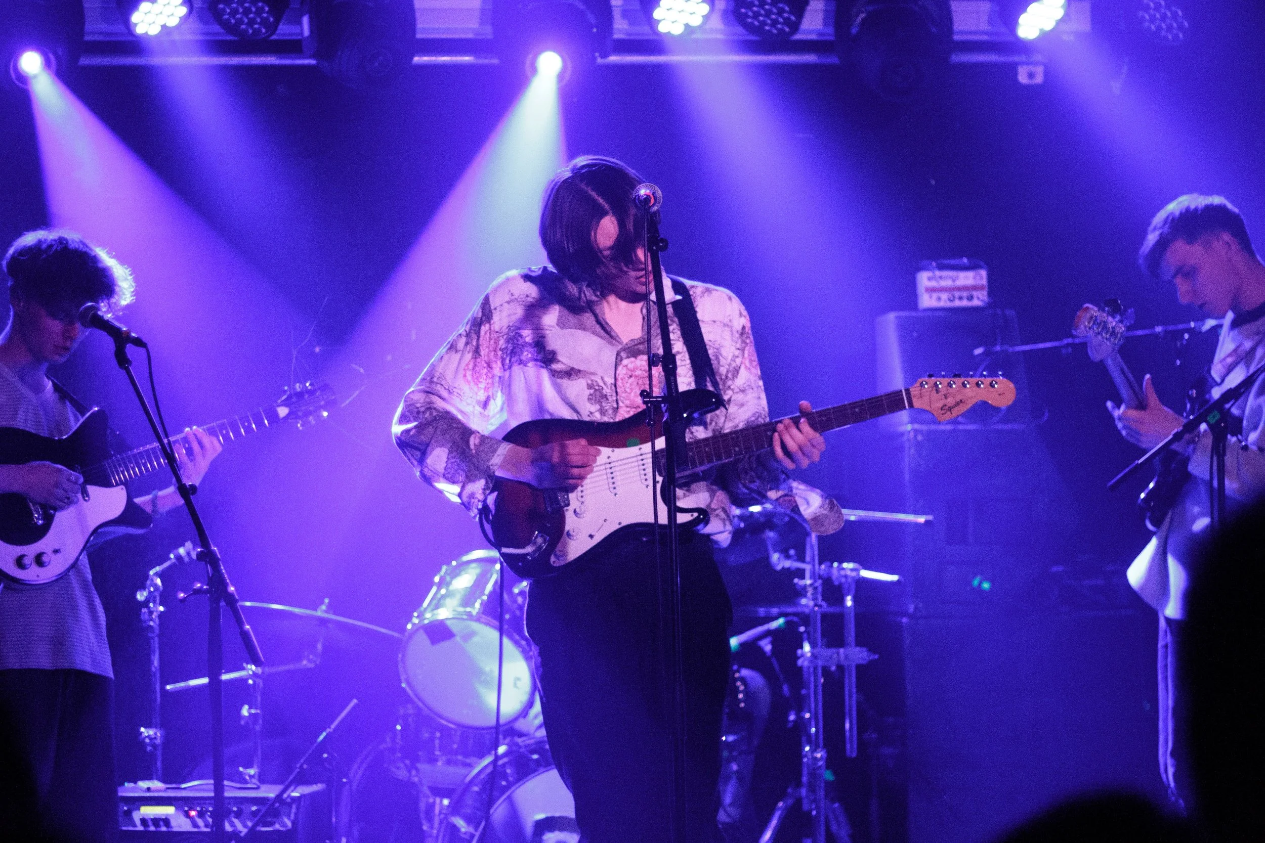 A band of three young men playing electric guitars on stage under purple and blue lighting, with a drum set in the background.