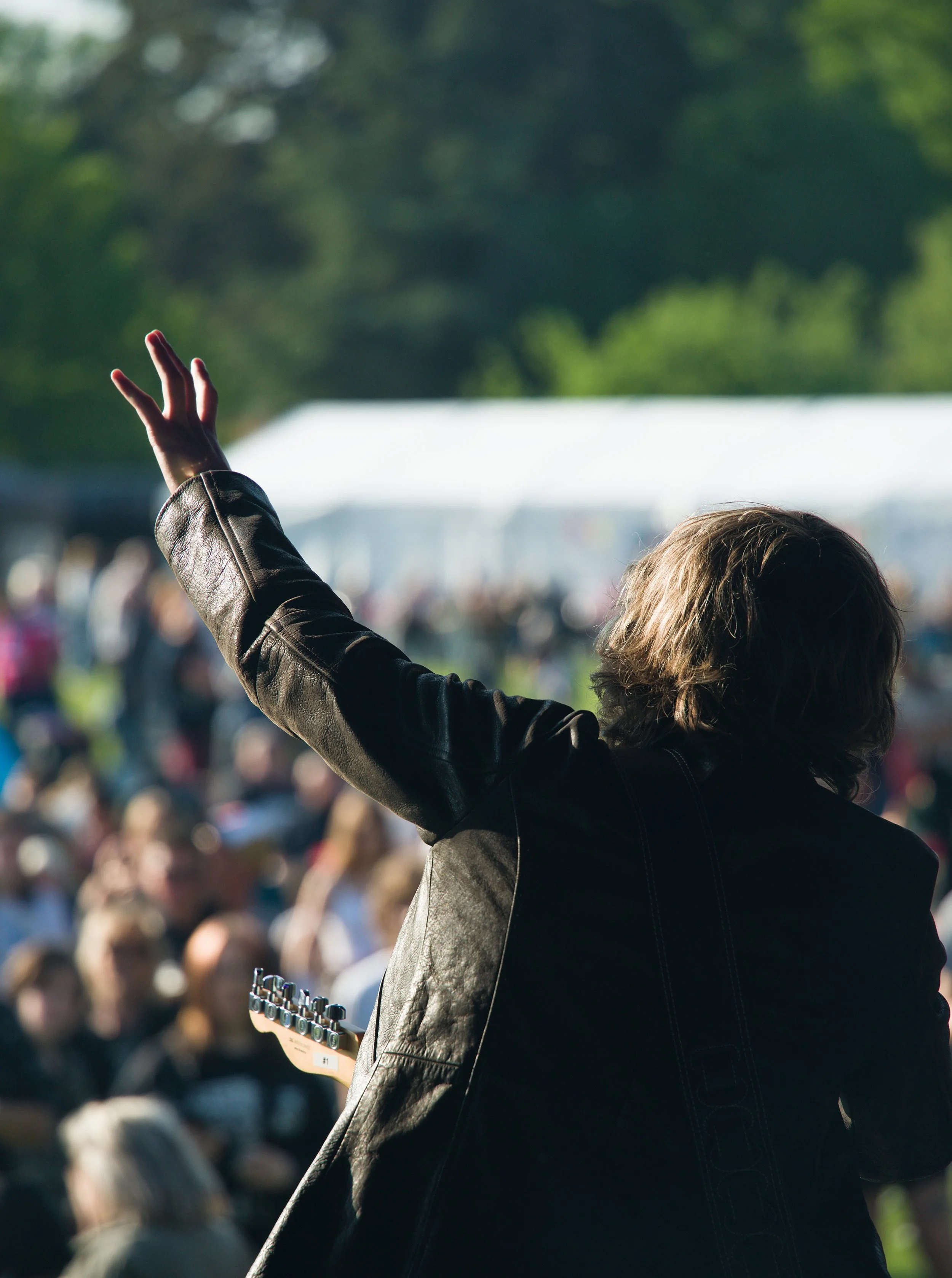 A person with shoulder-length brown hair, wearing a black leather jacket, raising their left hand while standing outdoors during daytime. The background shows a blurred crowd and trees.