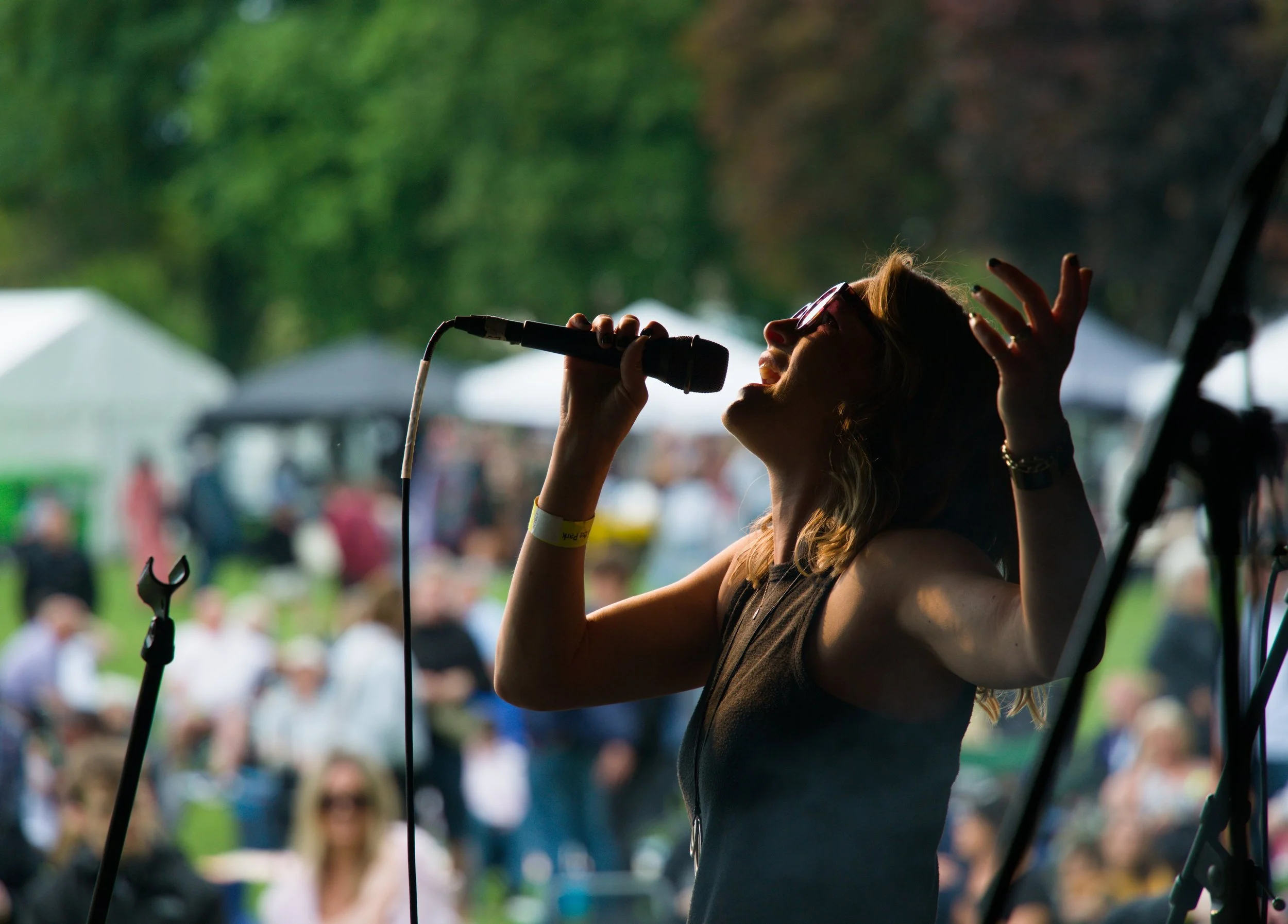 A woman holding a microphone singing at an outdoor event with a crowd in the background.