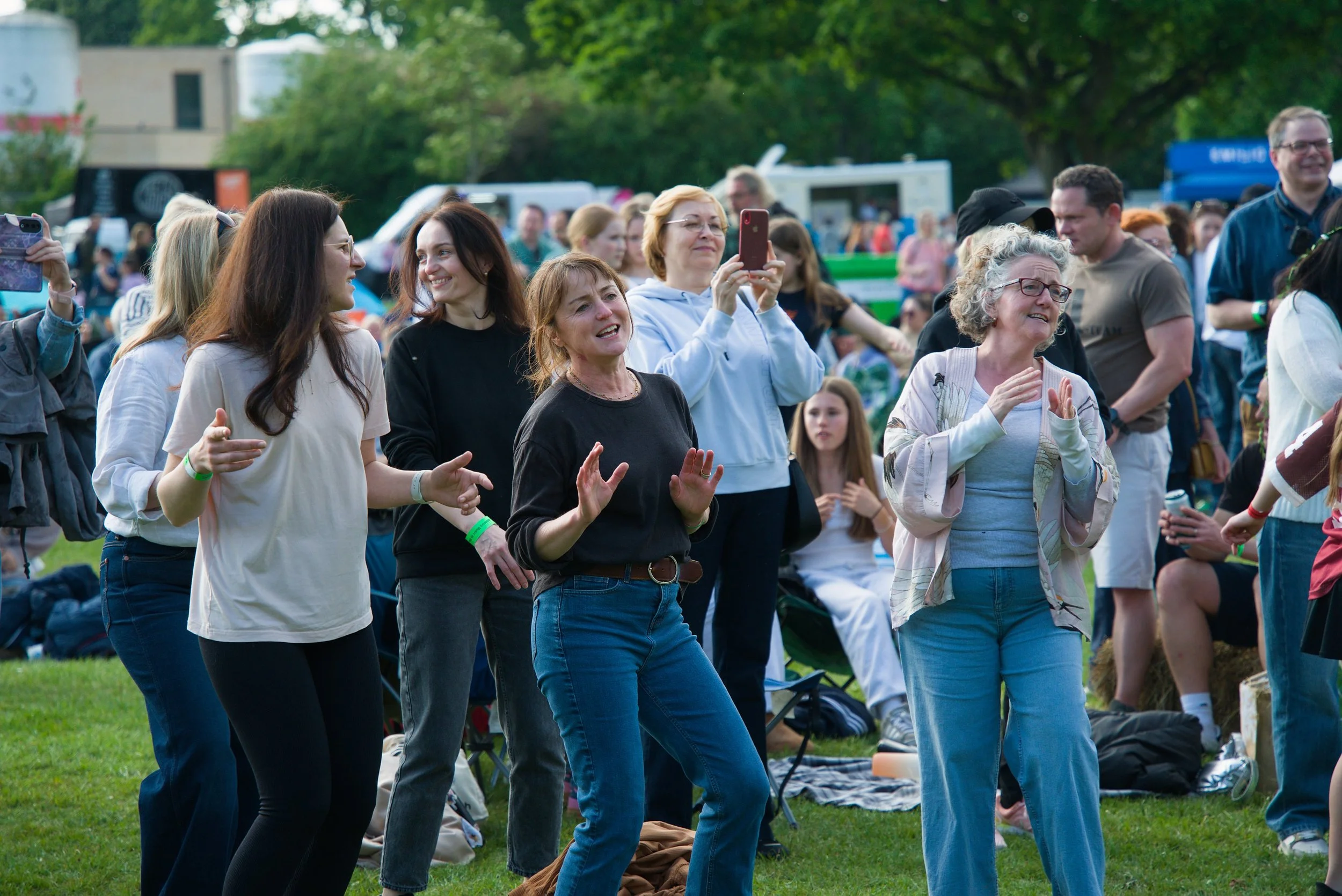 Group of people enjoying outdoor concert or event, standing and singing on a grassy area with trees in the background.