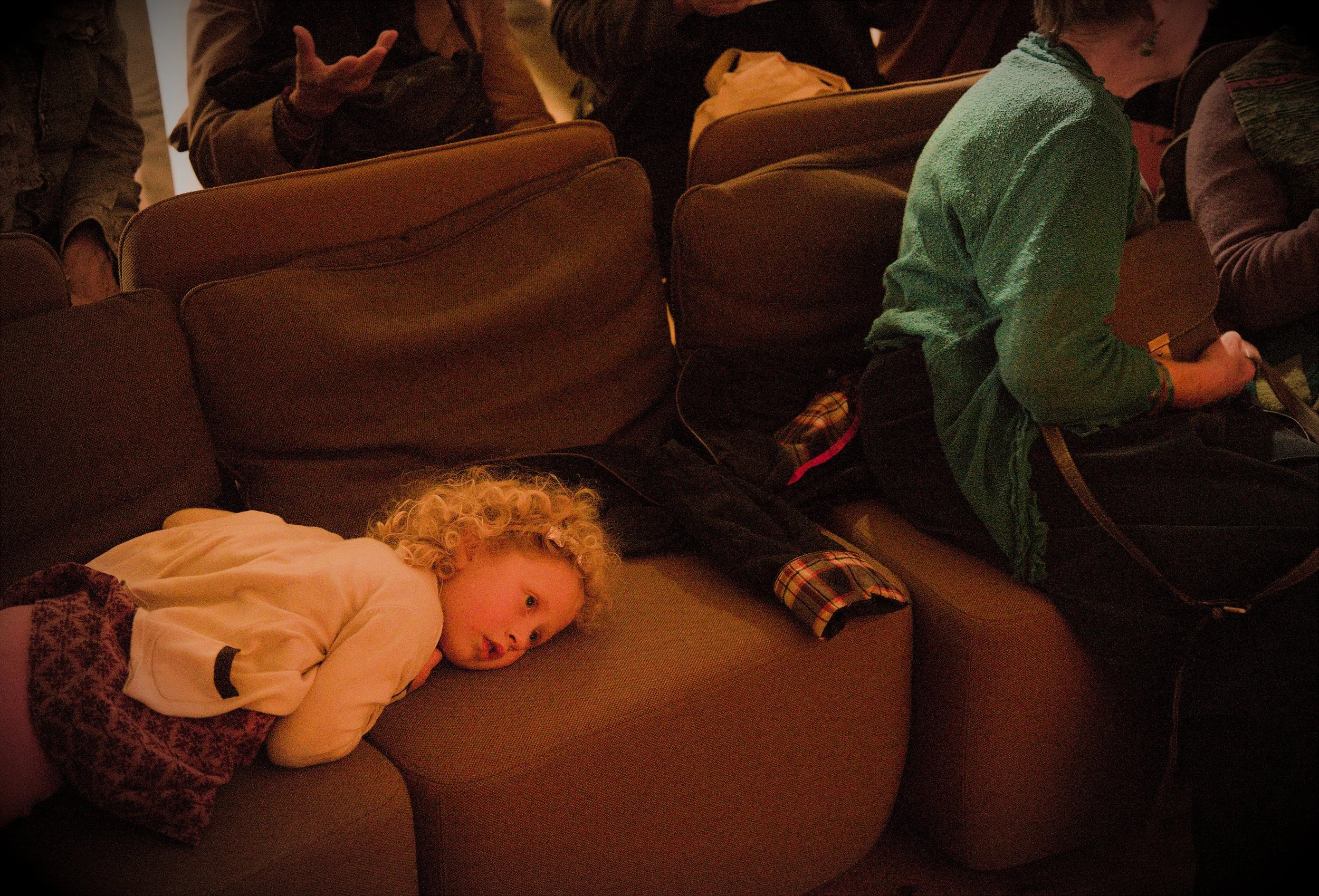 A young girl with curly blonde hair lying on a sofa, looking to the side, next to a woman sitting on the sofa in a green sweater, holding a phone. The scene appears to be indoors with dim lighting.