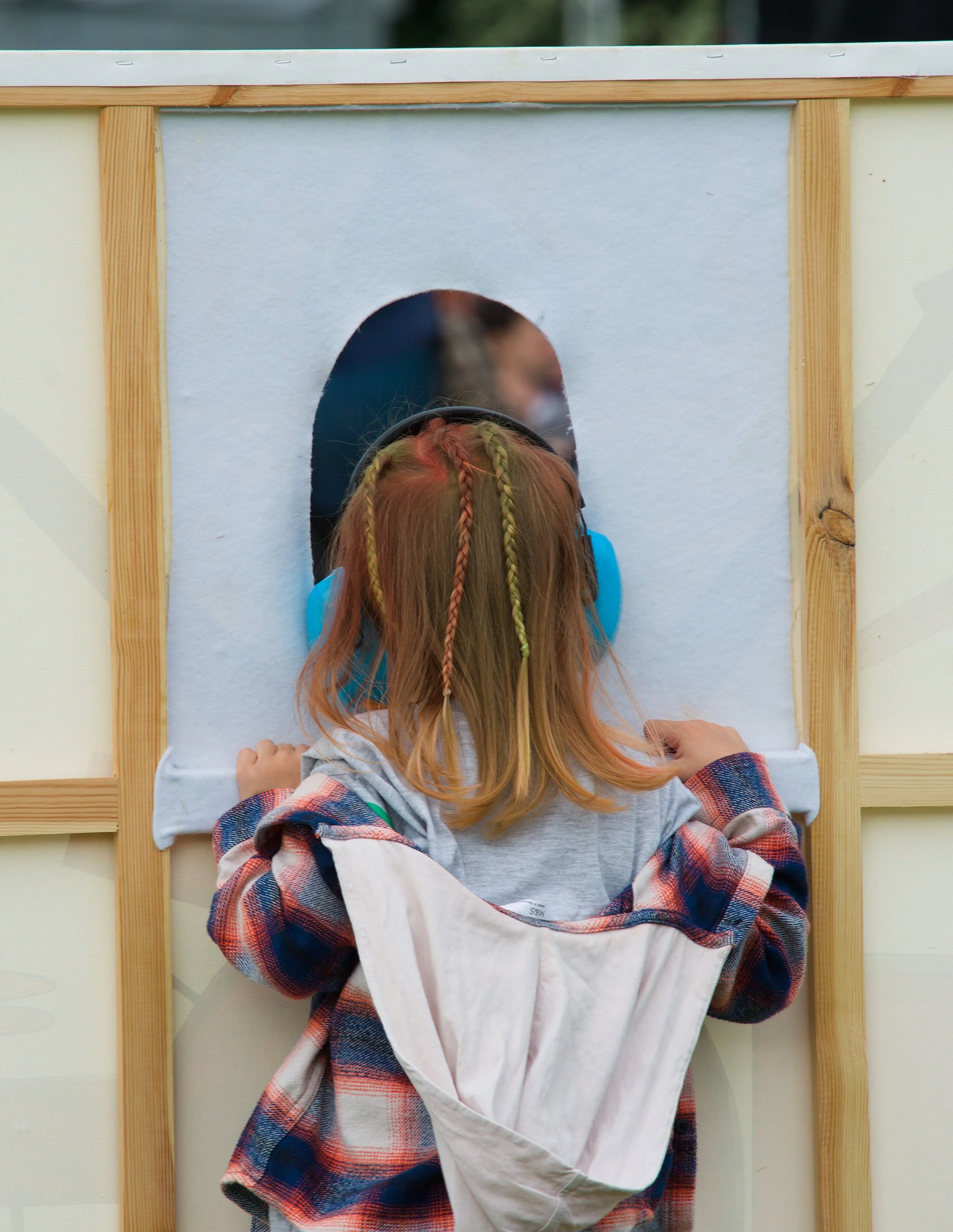 Child with long blonde hair in braids wearing a blue shirt, peeking through a small arched opening in a wooden structure, with hands on the bottom edge.