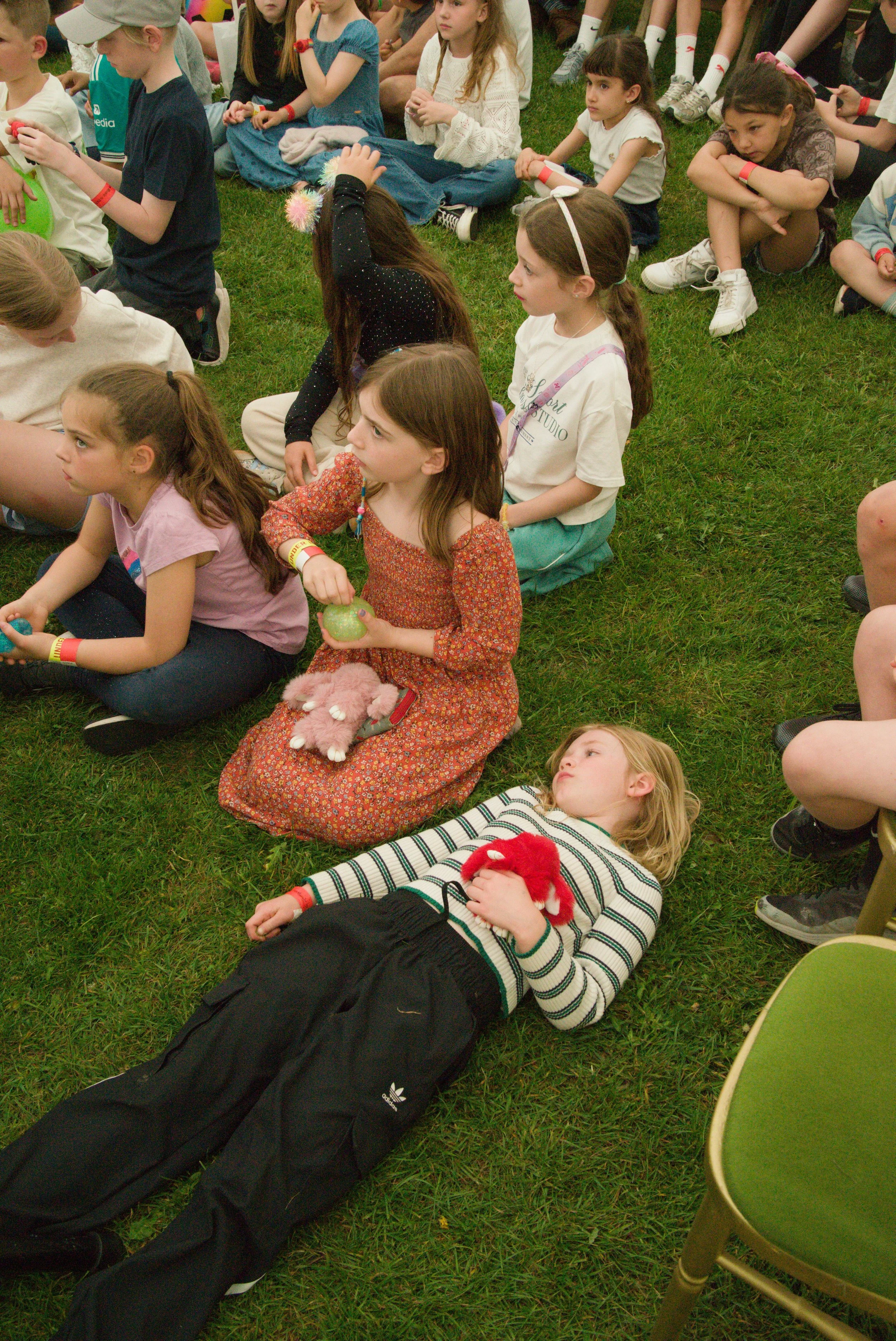 Children sitting on the grass during an outdoor event, some are listening attentively, and a girl is lying down holding a stuffed animal, with chairs visible nearby.