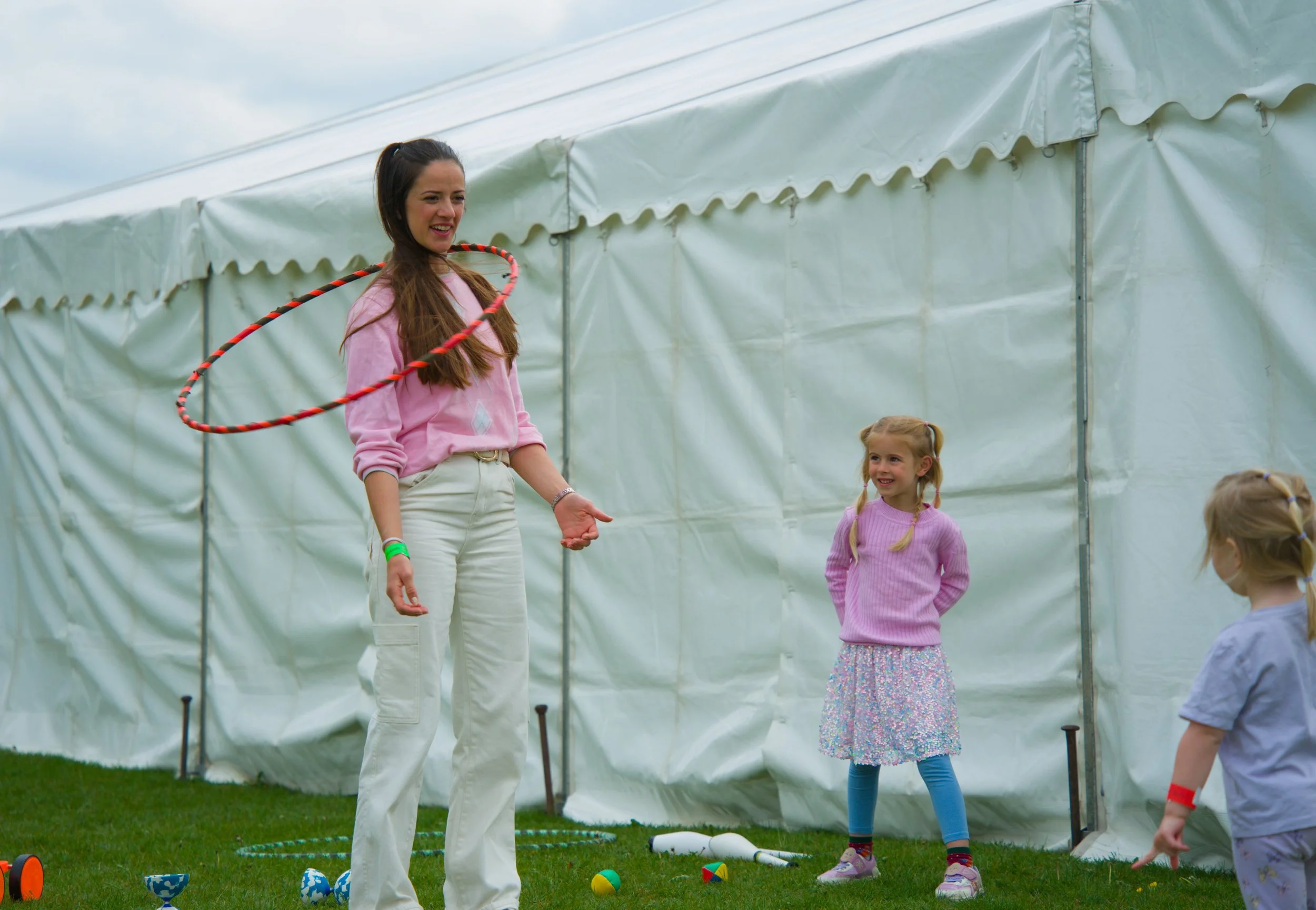 A woman with long brown hair hula hooping at a children's outdoor event near a white tent, with young girls and play equipment visible.