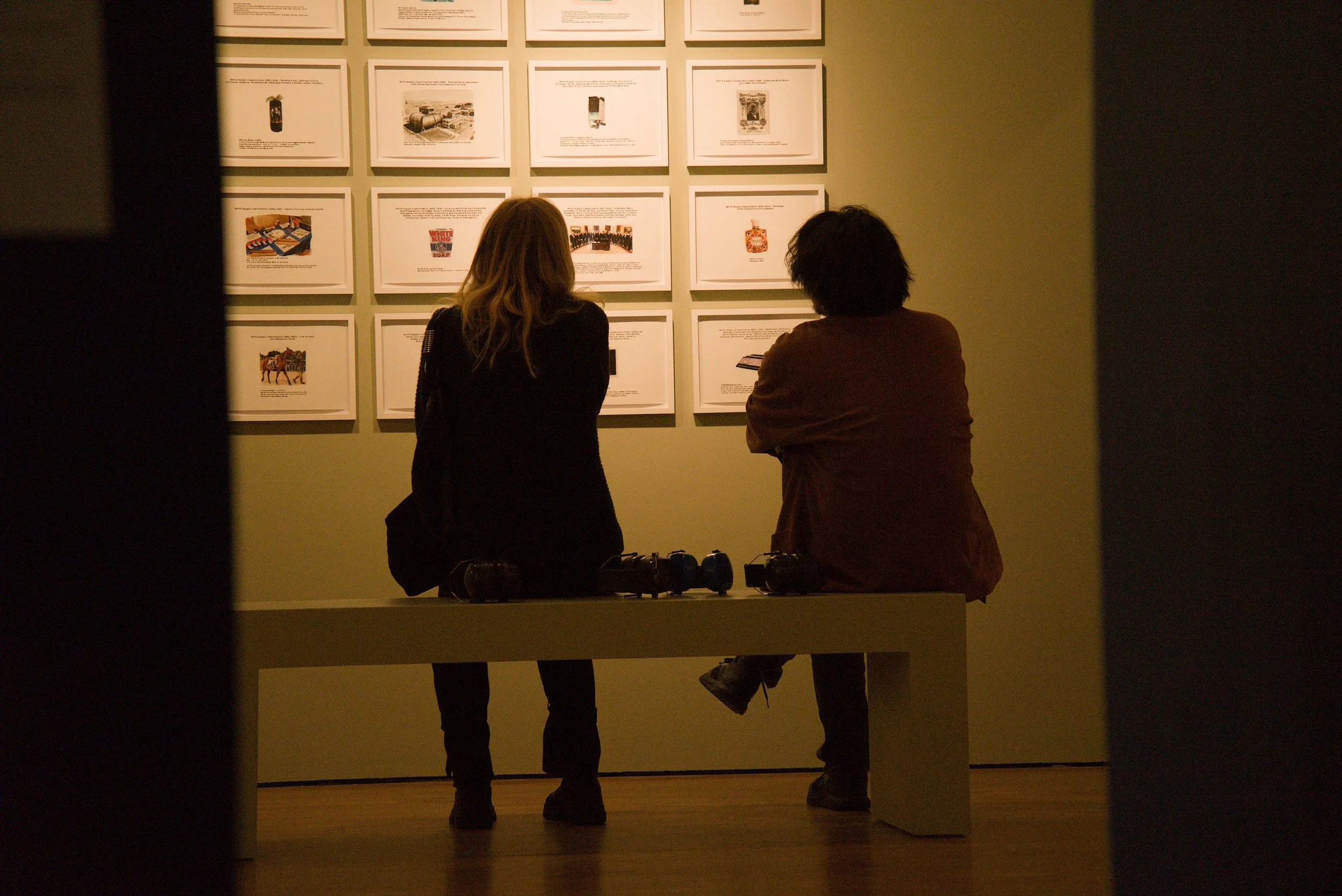 Two people observing framed pictures and text on a gallery wall. A table nearby holds several cameras.
