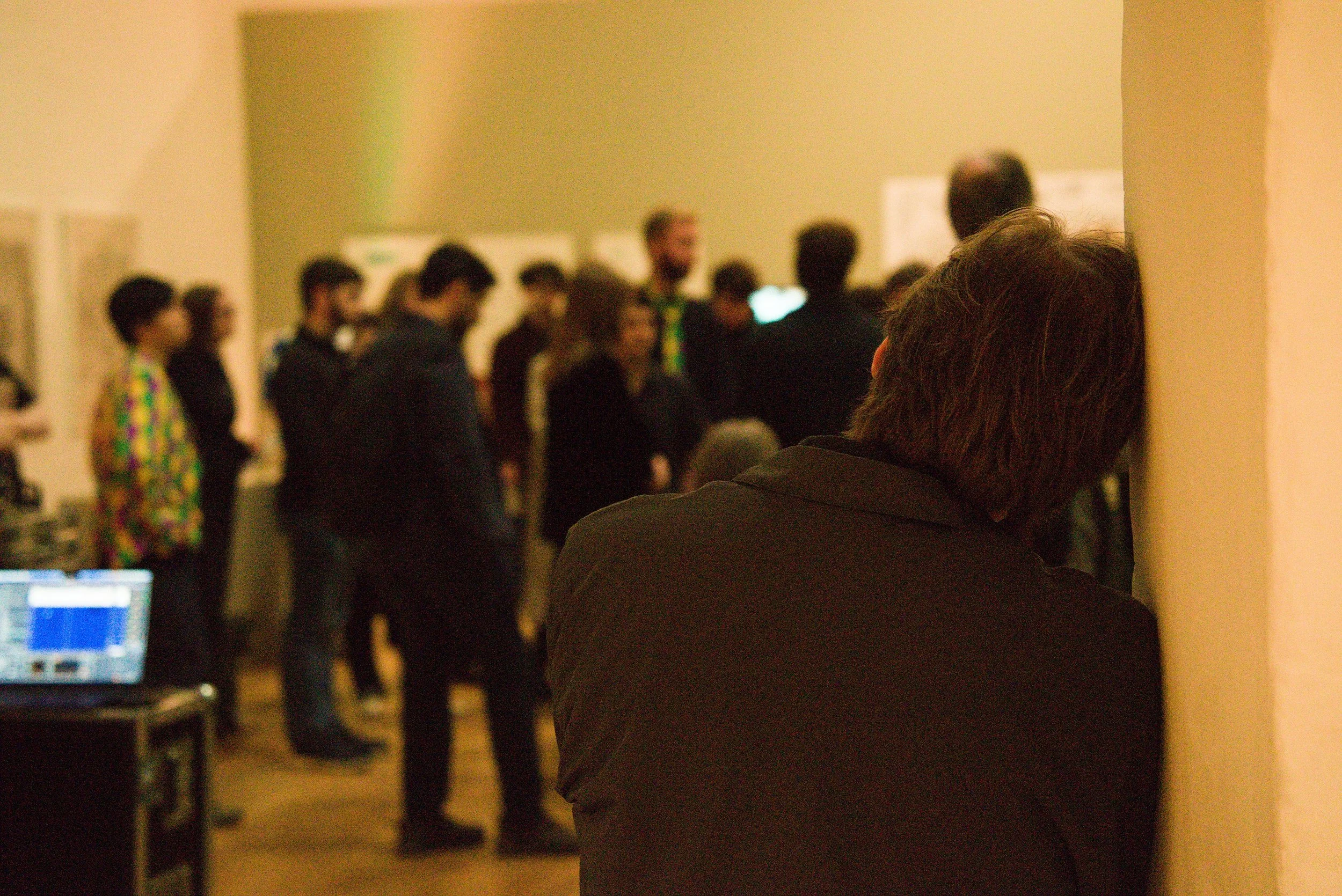 A person with dark hair and a black jacket is seen from behind, watching a group of people standing in a line in a well-lit indoor room, possibly at a presentation or event.