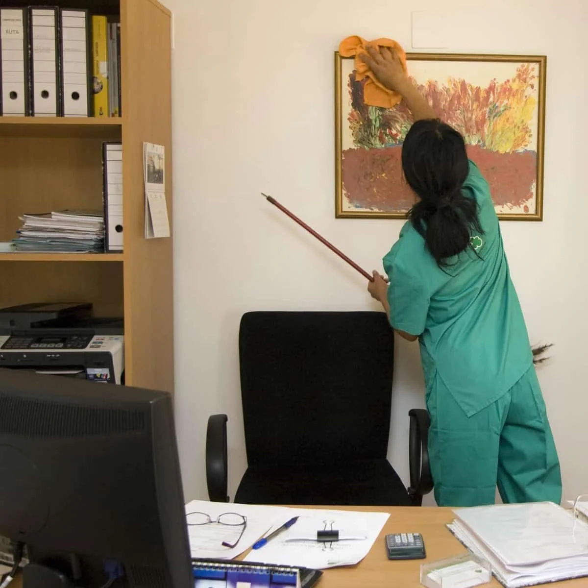 A person cleaning a framed picture on an office wall with an orange cloth, standing next to a black office chair and a desk with papers, glasses, a calculator, and a pen. There is a computer monitor and a bookshelf with binders and documents nearby.