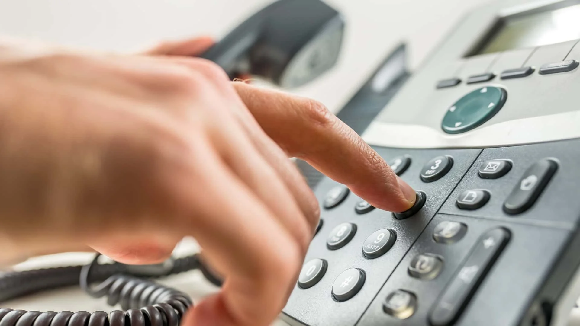 Close-up of a person's hand dialing a number on a black office telephone with a cord, silver buttons, and a digital display.