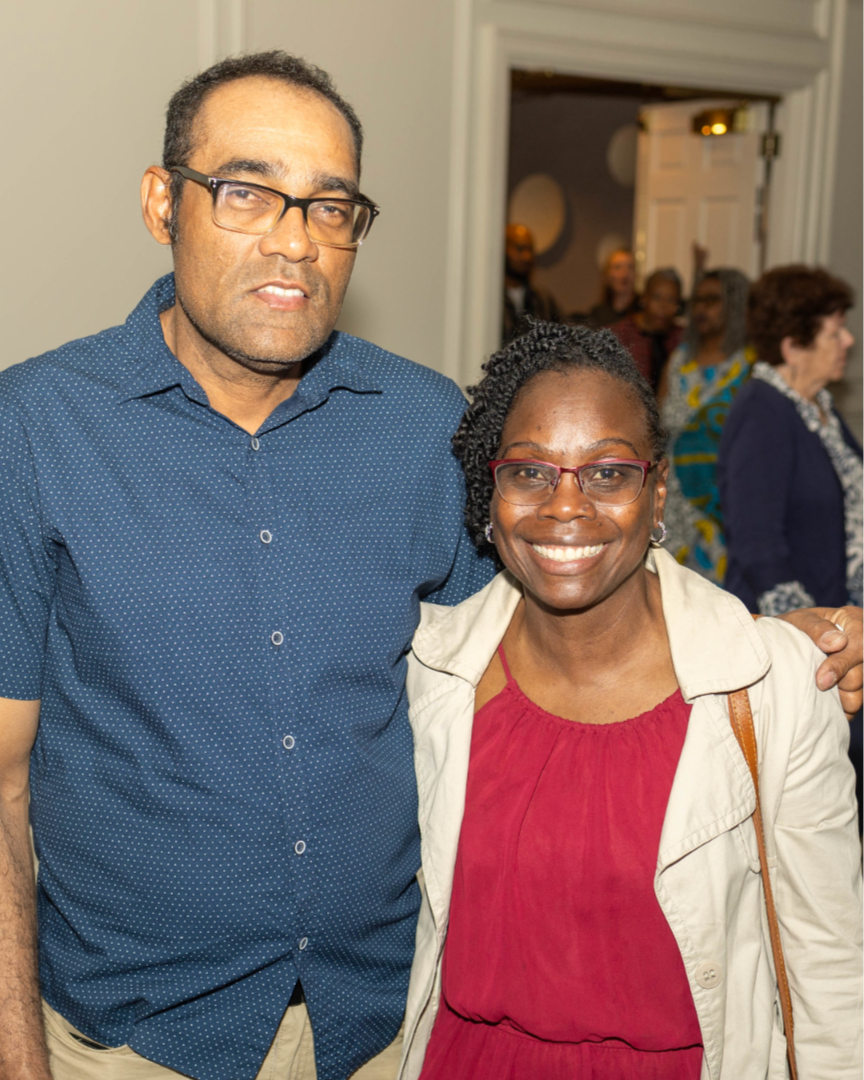 A man and woman smiling, with a crowd in the background, indoors at a social gathering.