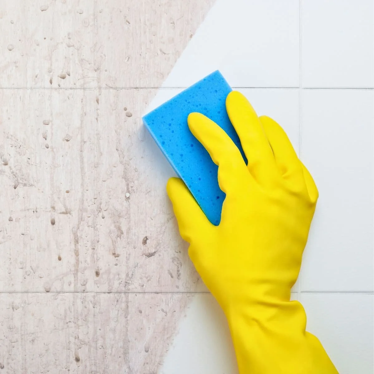 A person wearing a yellow rubber glove holding a blue sponge, cleaning white ceramic tiles on a wall.