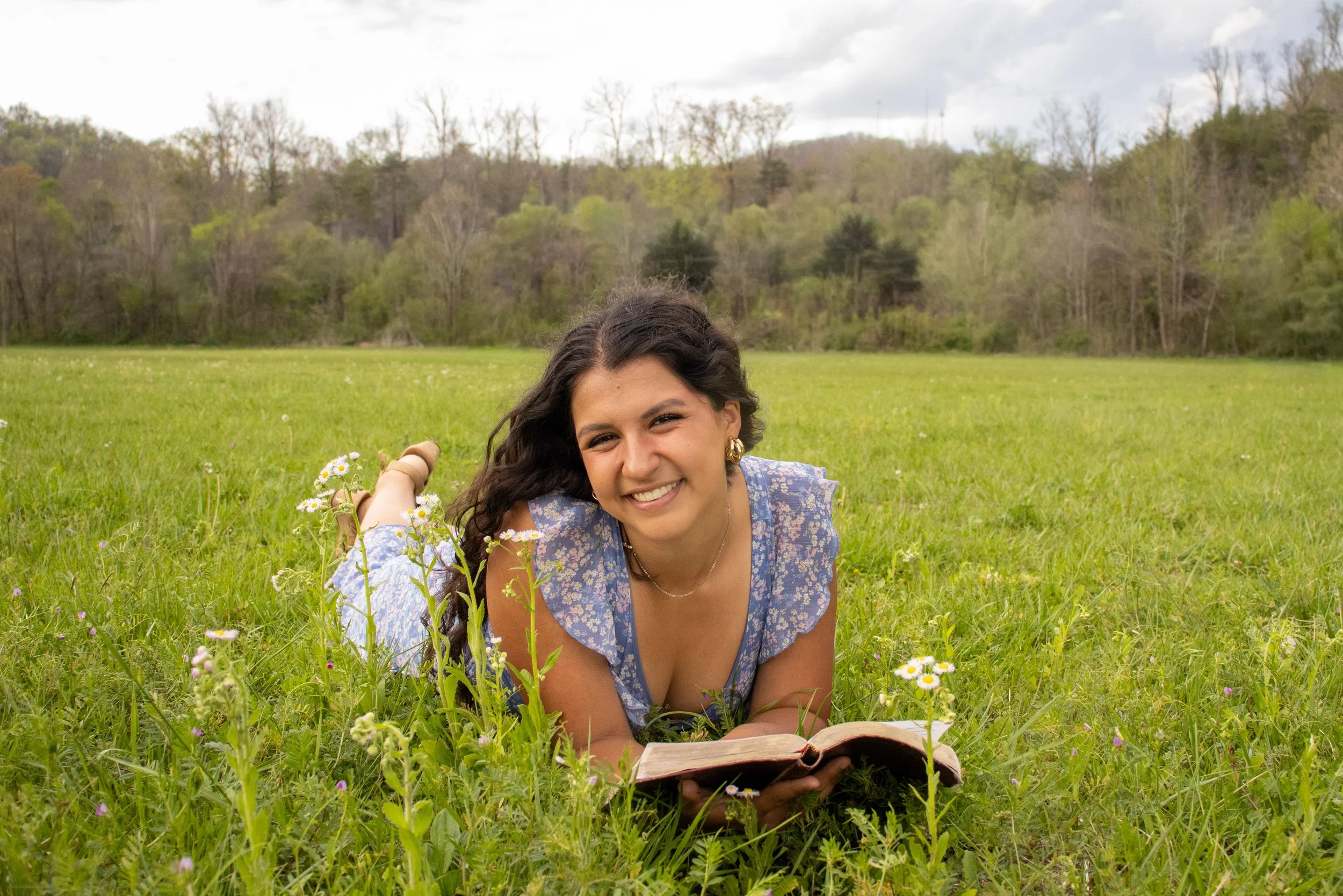A woman with dark, wavy hair lying on her stomach in a green grassy field with small white flowers, smiling at the camera while holding an open book, with trees and cloudy sky in the background. Senior Photo, Graduation