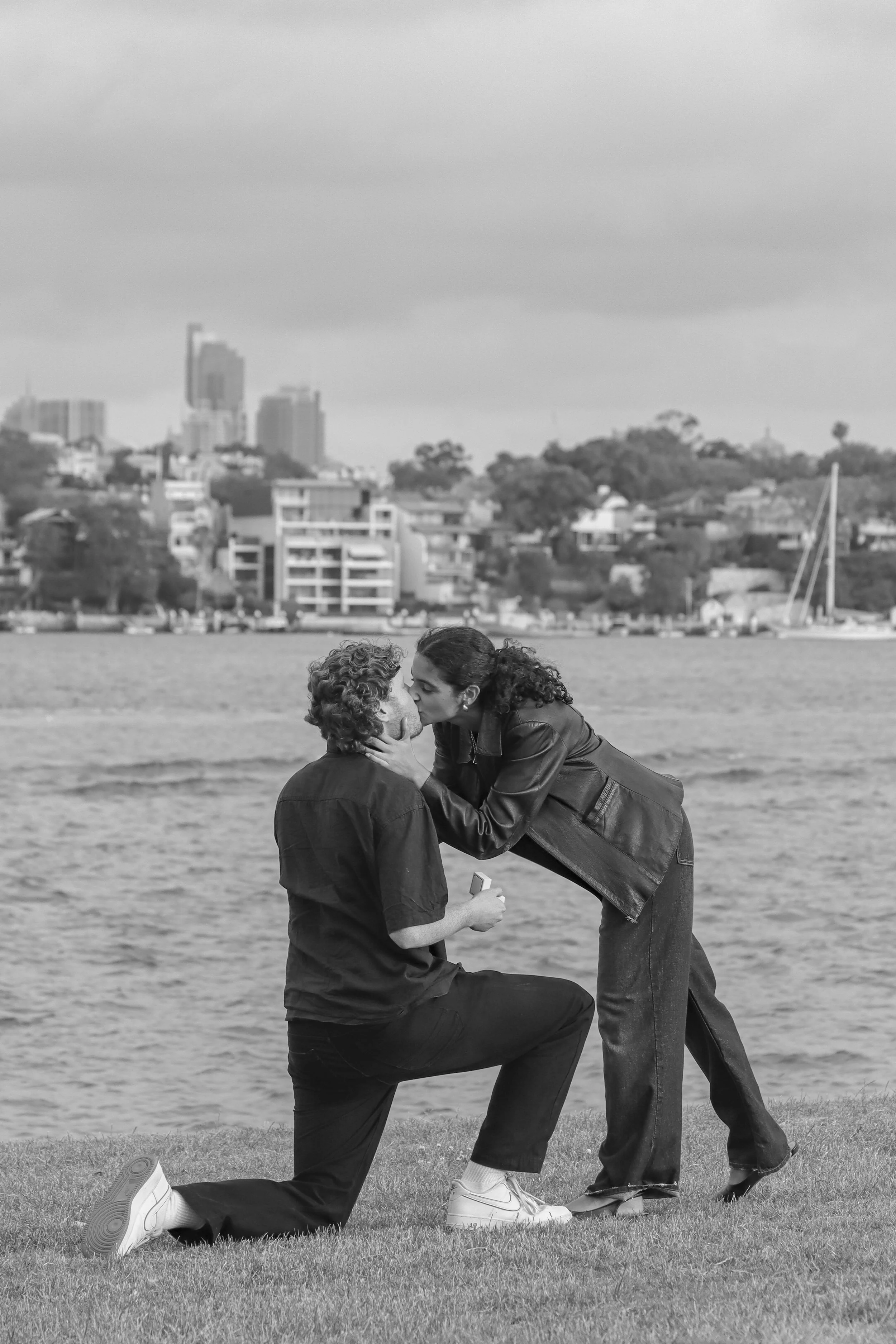 A black-and-white photo of a couple kissing outdoors near a body of water with a city skyline and hills in the background. sydney australia, engagement photos