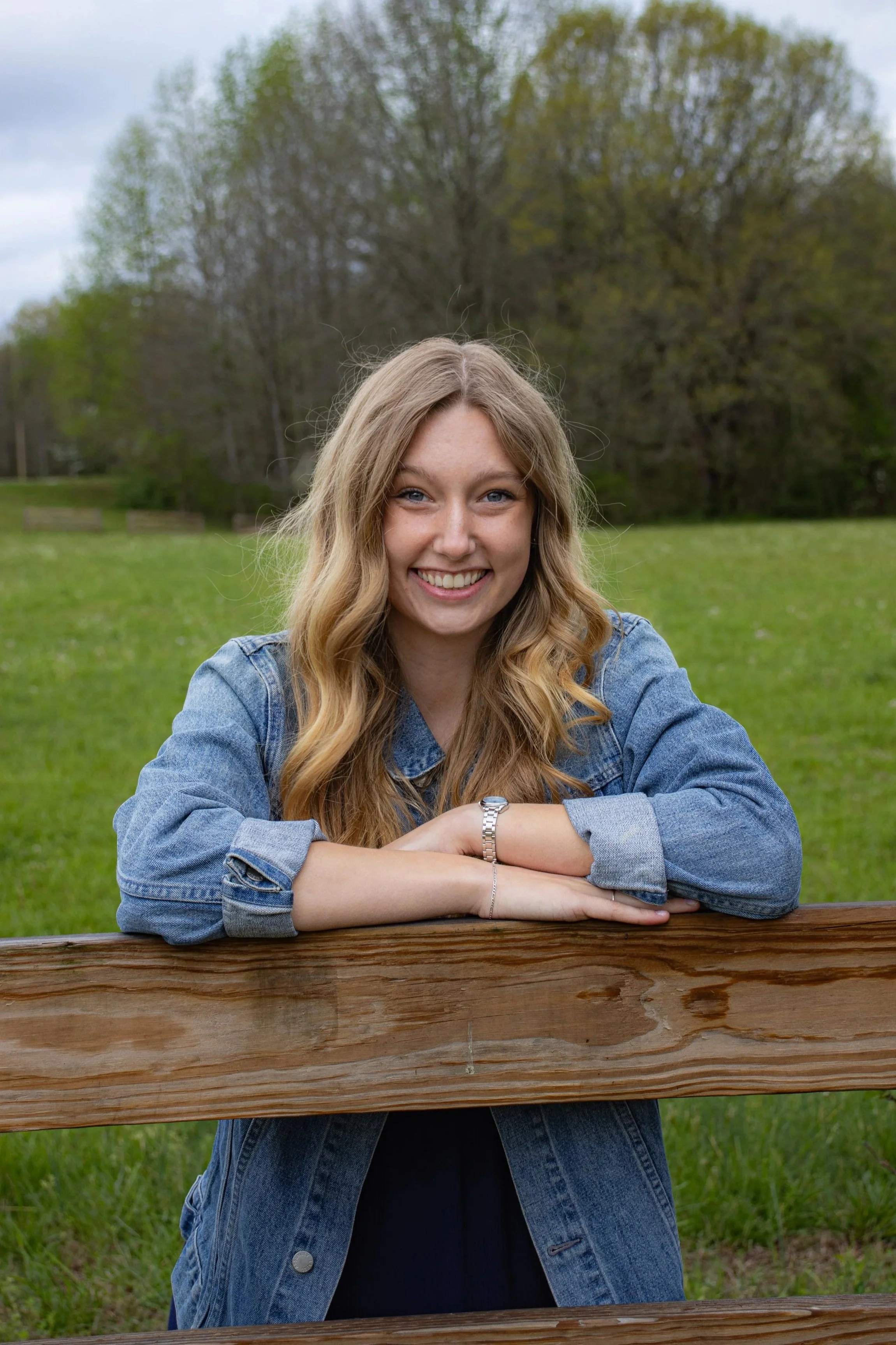 A young woman with long blonde hair smiling, leaning on a wooden fence in a park with trees and grass in the background. Danielle Brower