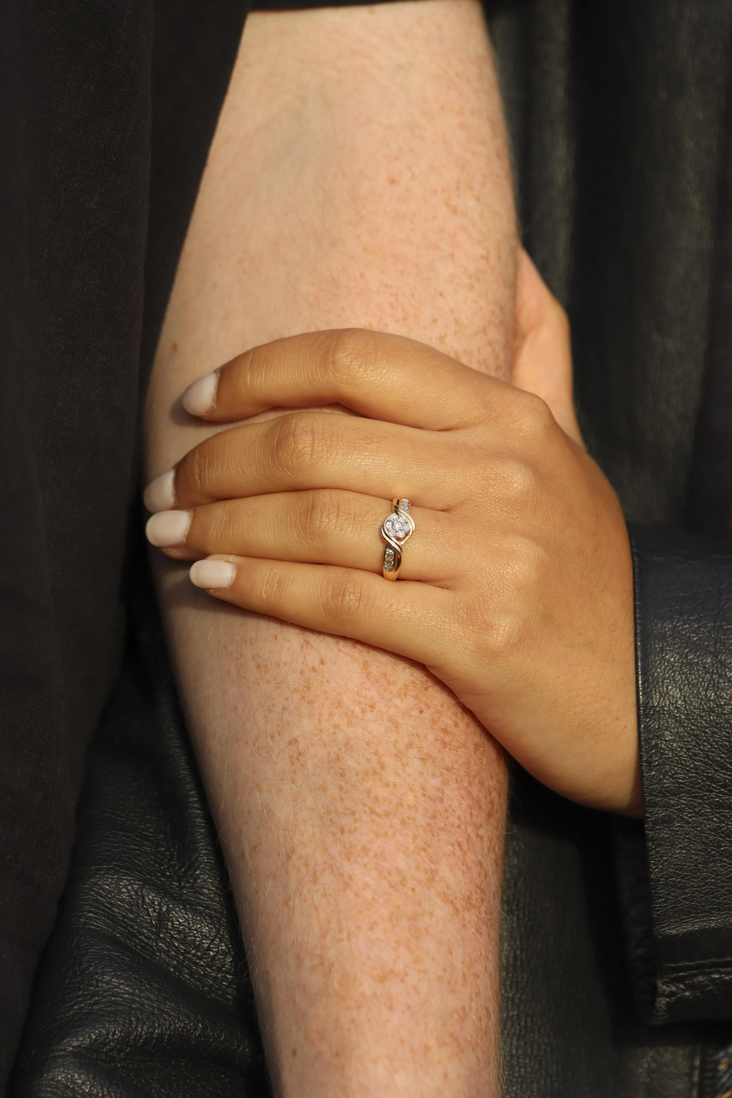 A woman wearing a diamond ring on her finger, resting her hand on mans arm with freckles, against a black leather and dark fabric background. engagement photos