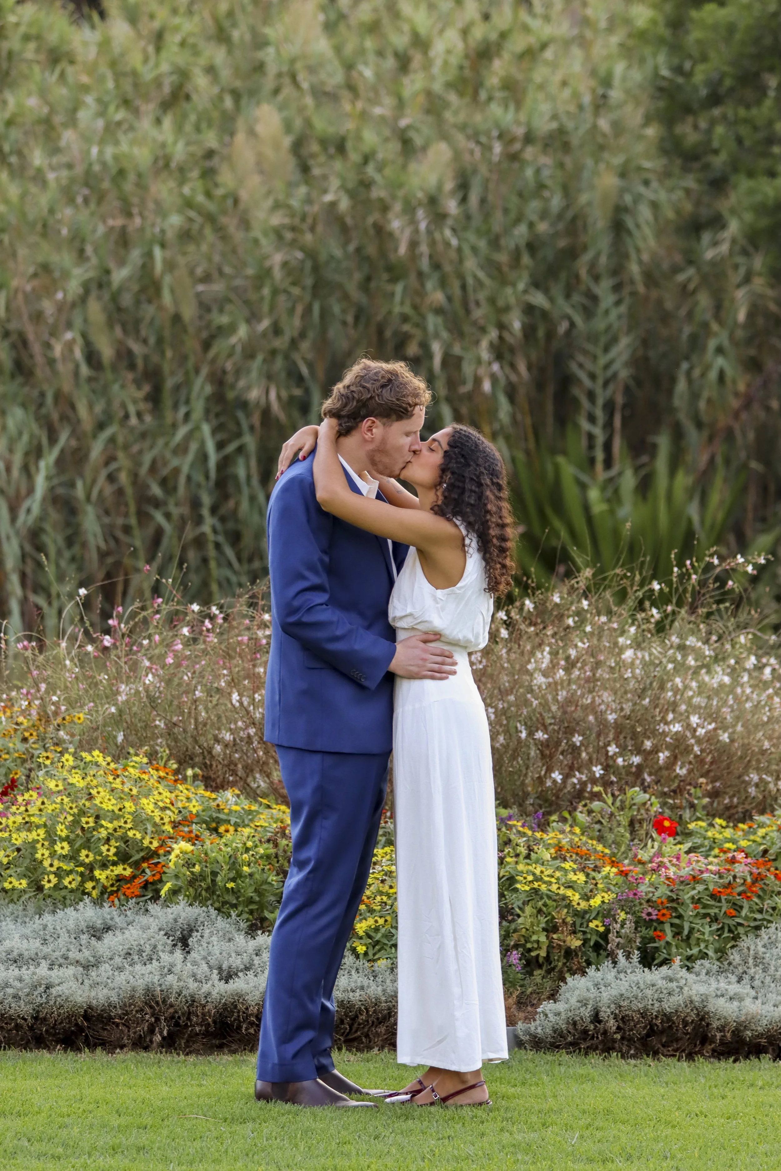 A man in a blue suit and a woman in a white dress are kissing outdoors, surrounded by colorful flowers and tall greenery. Wedding photos