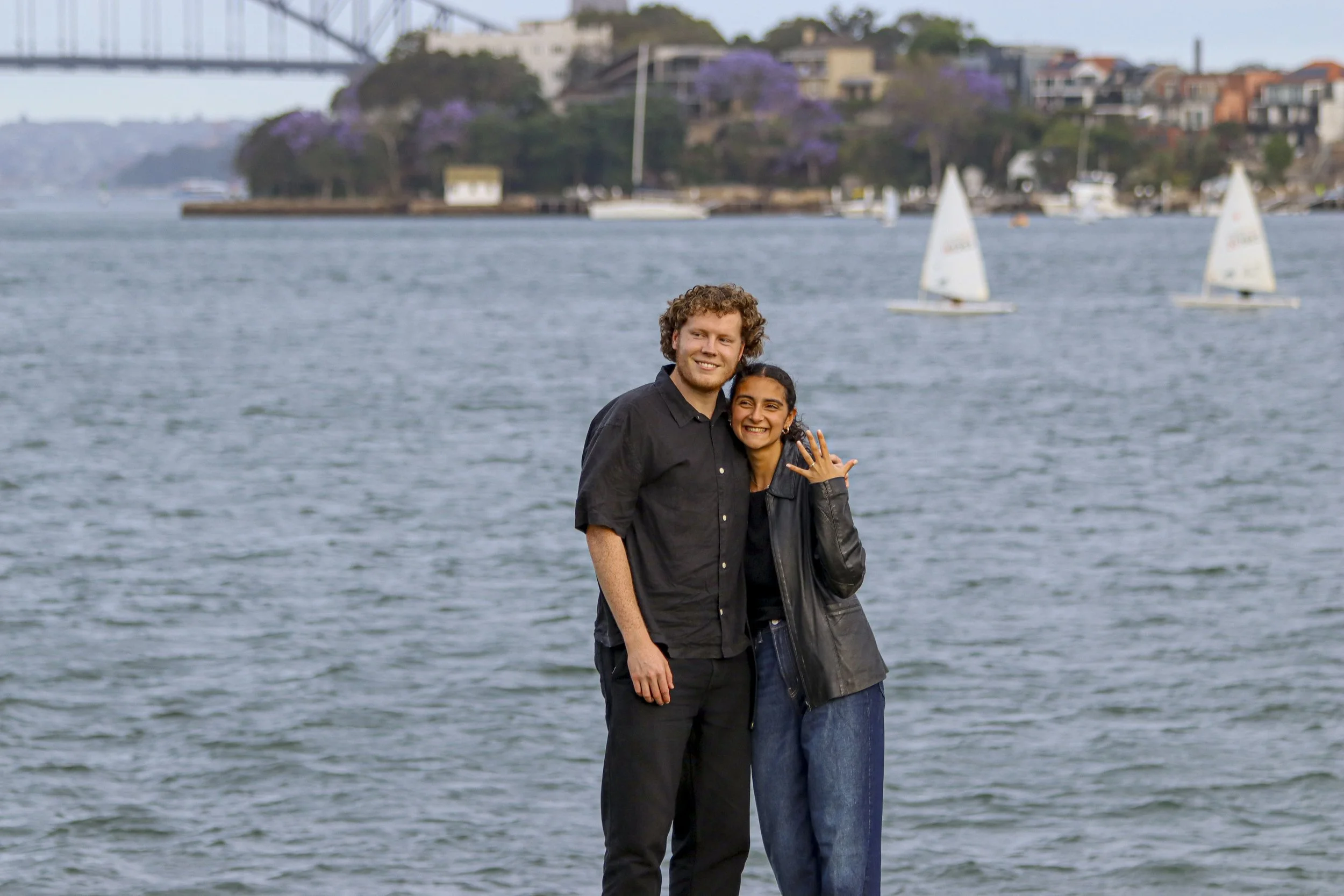 A man and a woman standing by a body of water, smiling and posing for a photo with sailboats in the background. The woman is showing off a ring on her hand. engagement photos