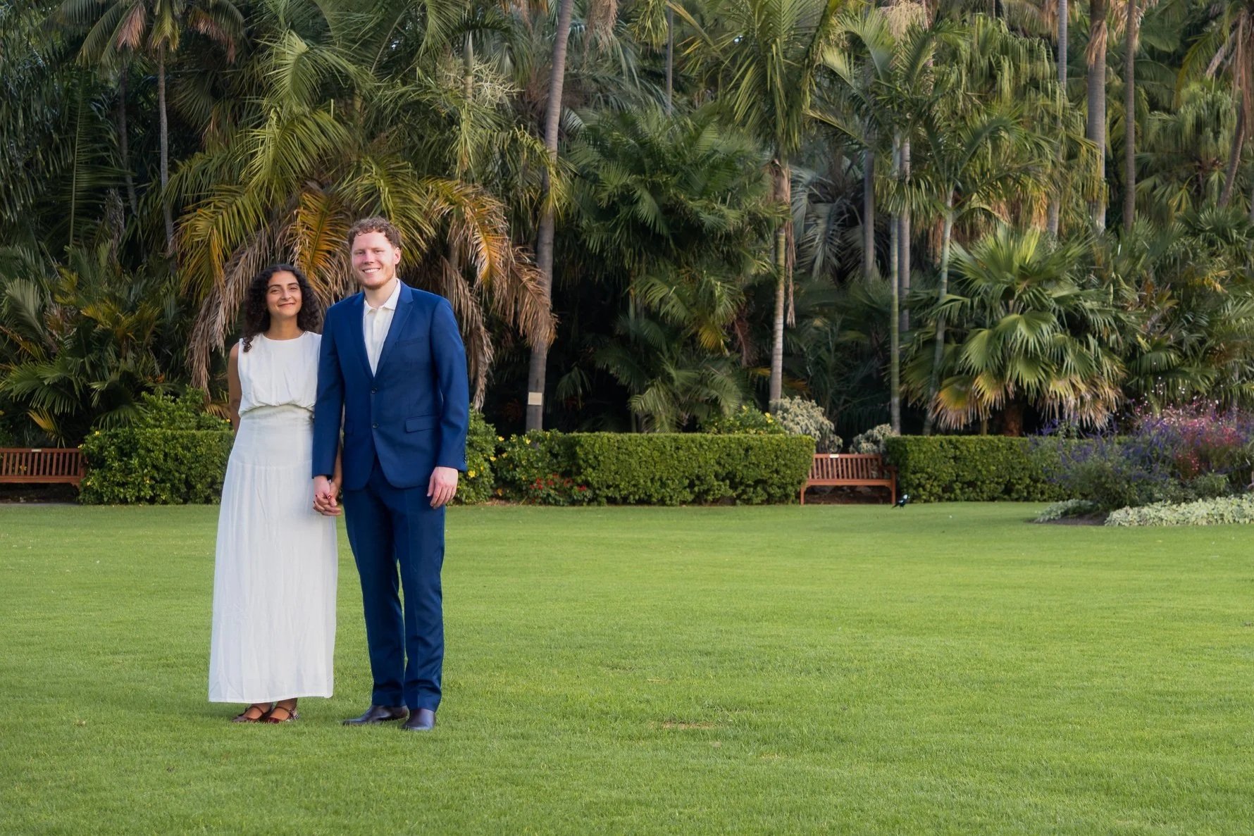 A couple standing hand in hand on a well-manicured lawn in a garden with tall green palm trees and bushes in the background. Wedding Photo