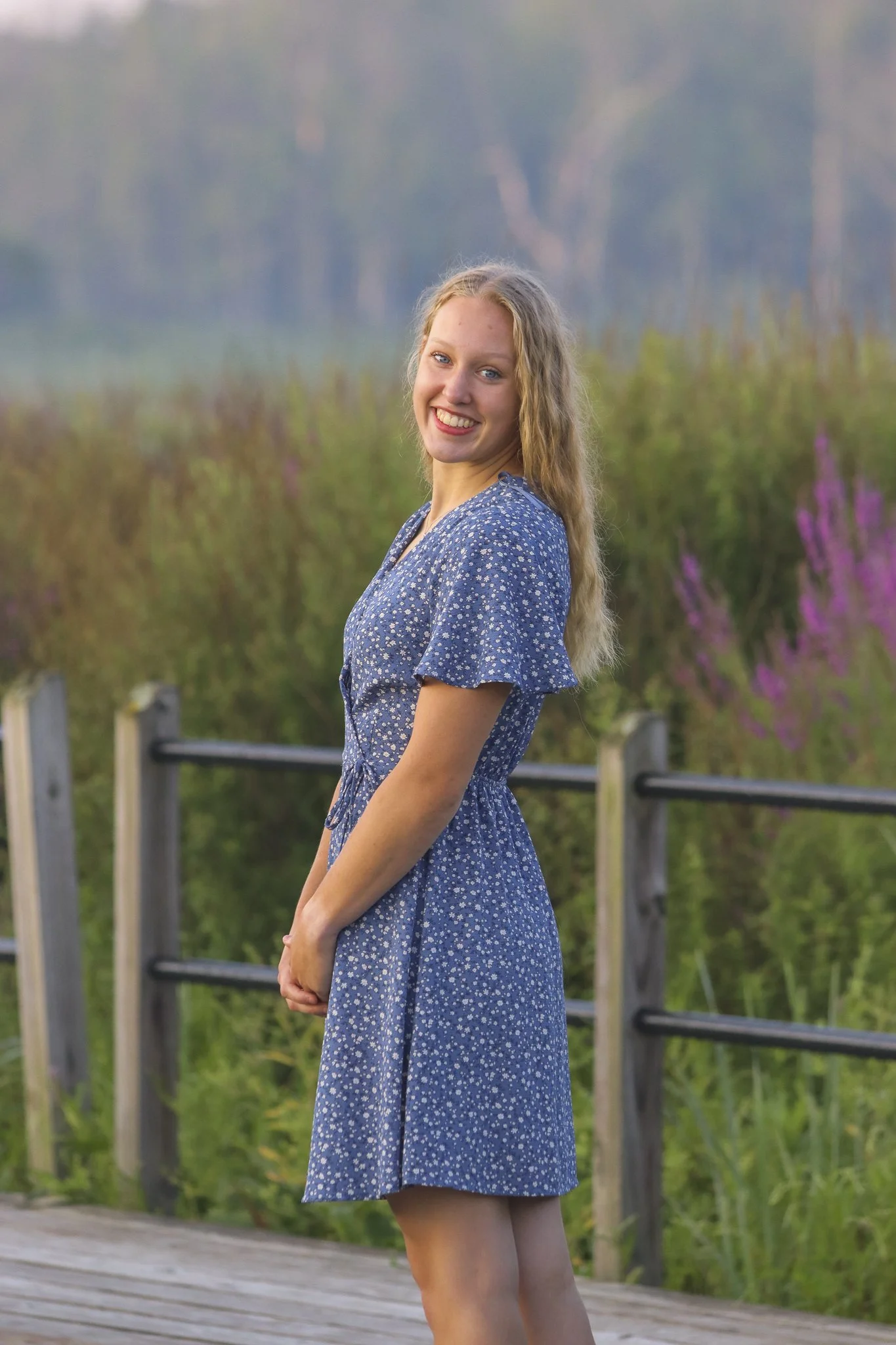A young woman with long, curly blonde hair smiles and looks at the camera while standing outdoors on a wooden pathway, wearing a blue dress with small white floral patterns. The background features lush greenery and flowering bushes. Senior Photo,