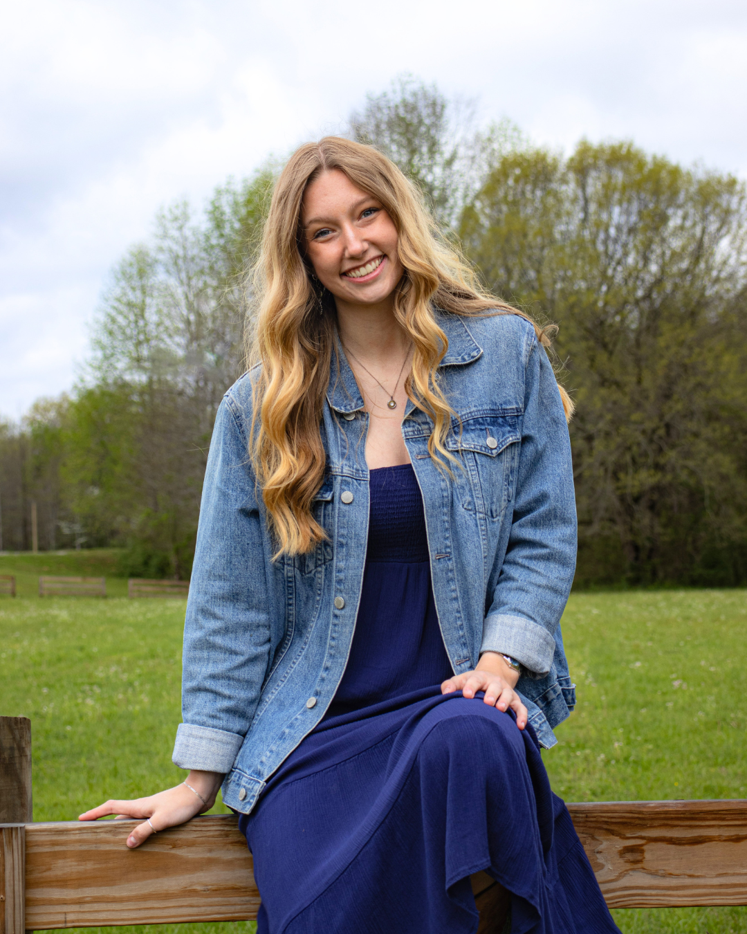 A young woman with long wavy blonde hair smiling while sitting on a wooden fence outdoors, wearing a blue dress and denim jacket, with trees and a cloudy sky in the background.