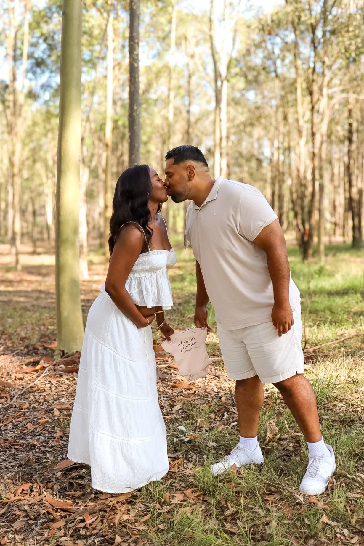 A couple kissing in a forest, holding a small baby's outfit. The woman is wearing a white dress, and the man is in a beige shirt and shorts. Pregnancy announcement