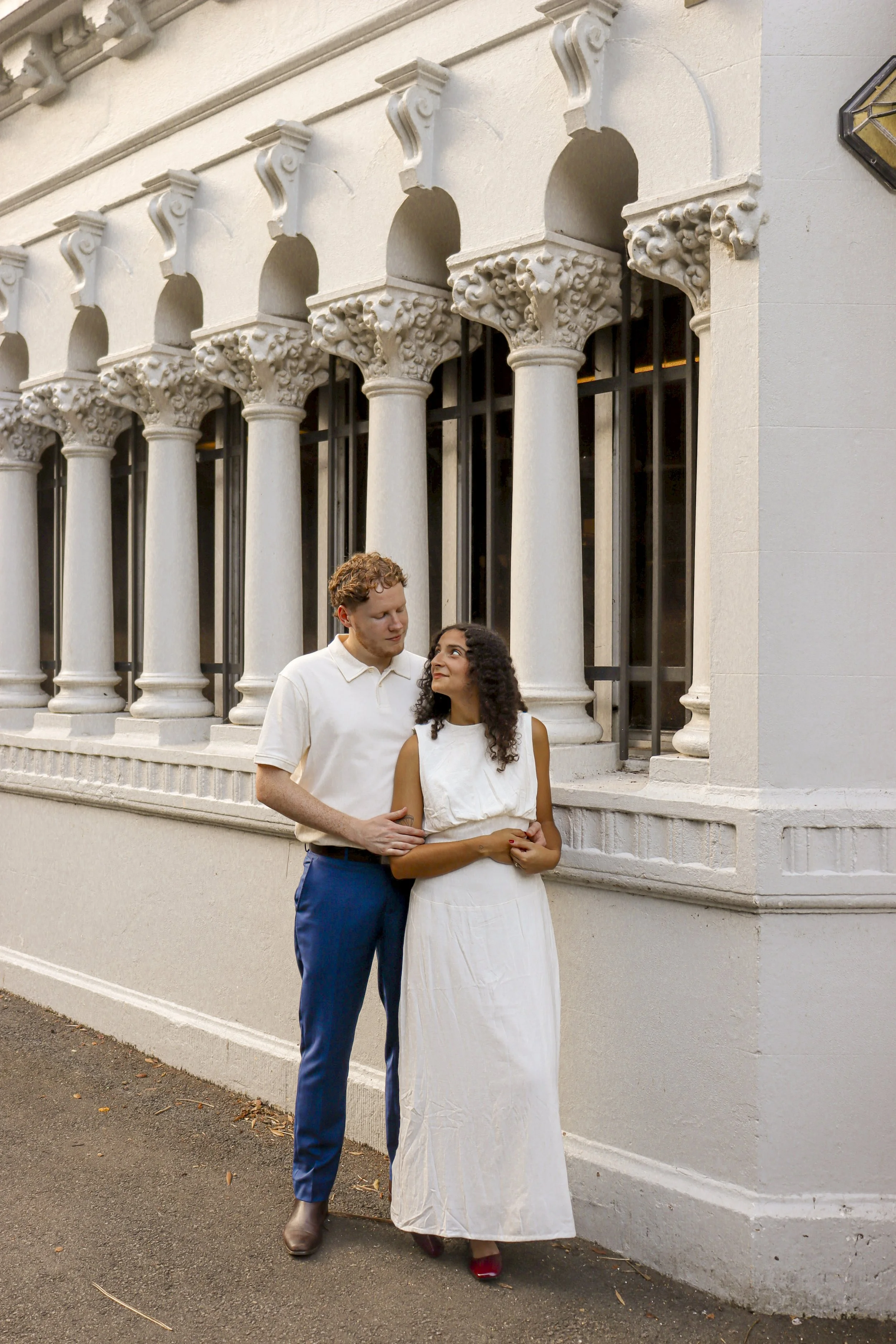 A couple standing close together outside a building with ornate white columns and arches. The man has curly red hair, wearing a white polo shirt and blue pants, while the woman has dark curly hair, wearing a sleeveless white dress and red shoes, looking at each other affectionately.