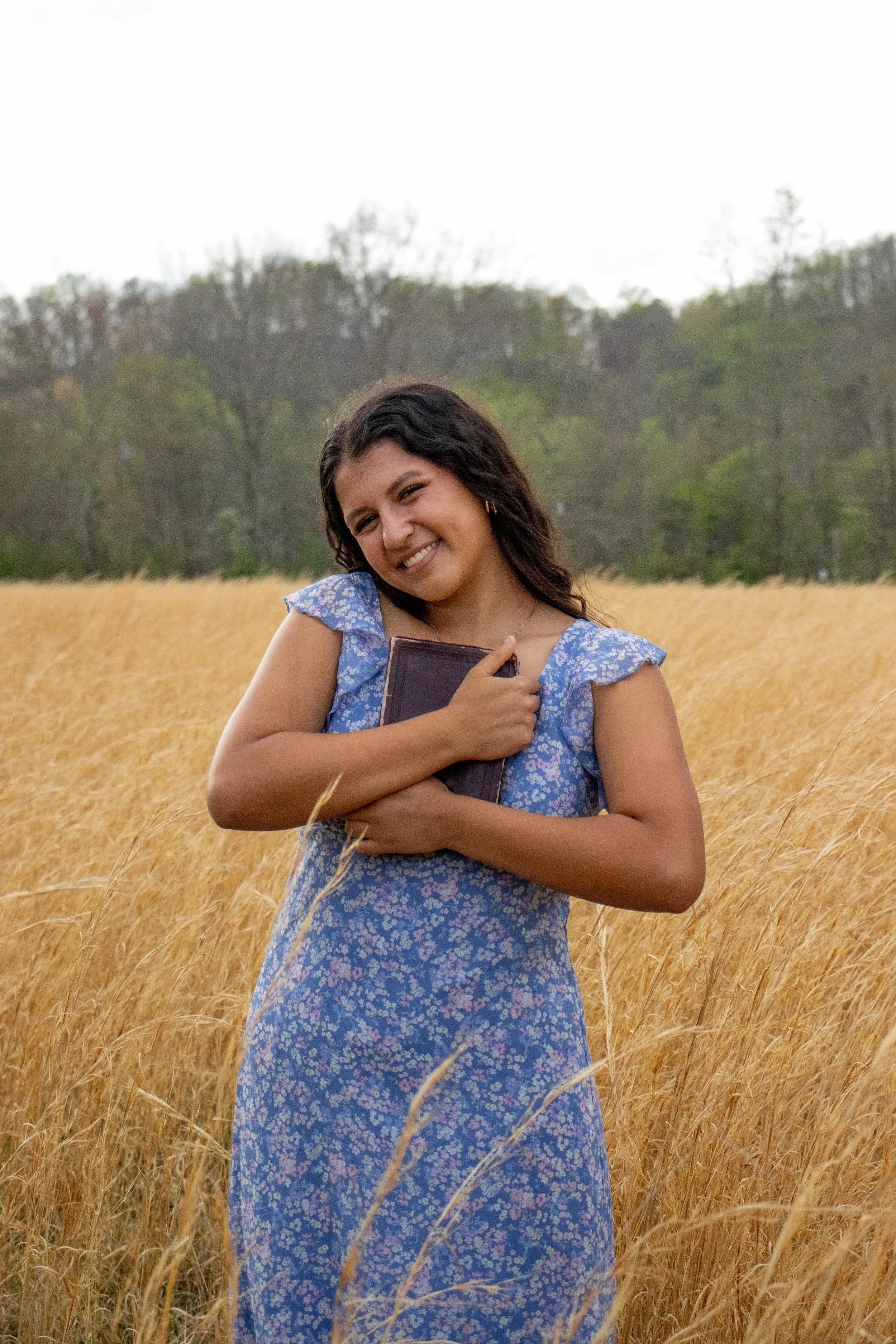 A smiling woman in a blue floral dress holding a book in a golden field of tall grass with trees in the background. Senior photos, graduation, outdoorsy