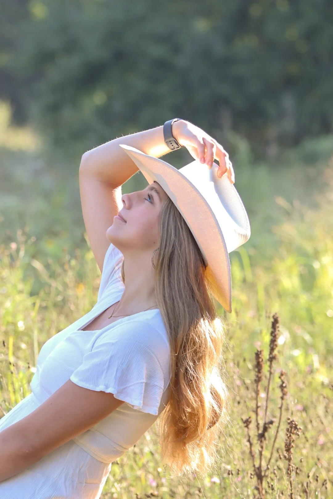 Young woman in a white dress and wide-brimmed hat standing in a sunlit field, smiling and looking upward. Cowboy, western, senior photo, graduation