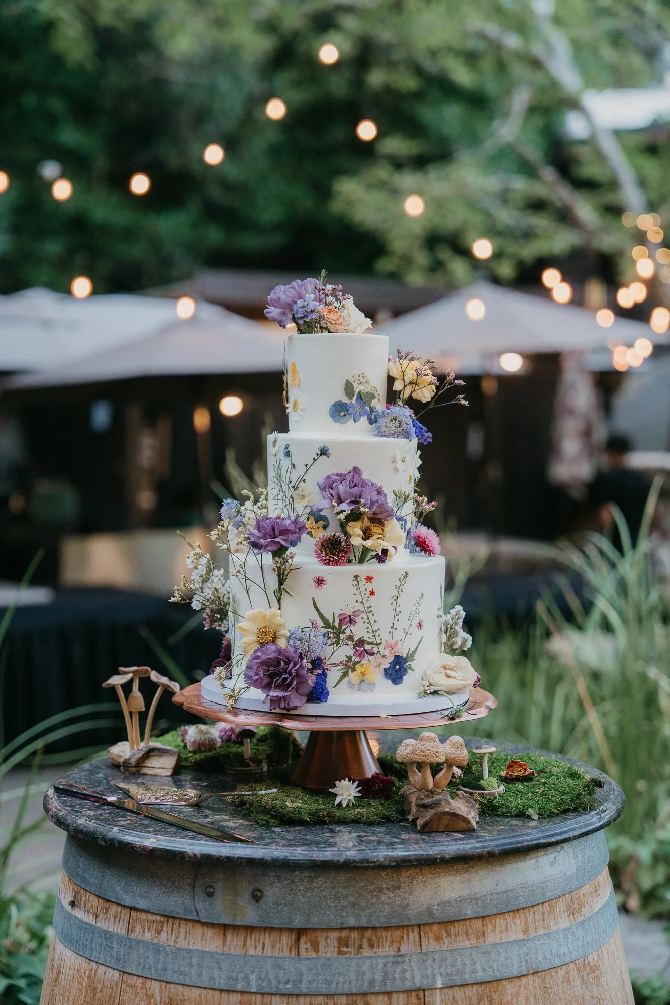 Three-tier wedding cake with pressed flowers under string lights at Wildwood Acres