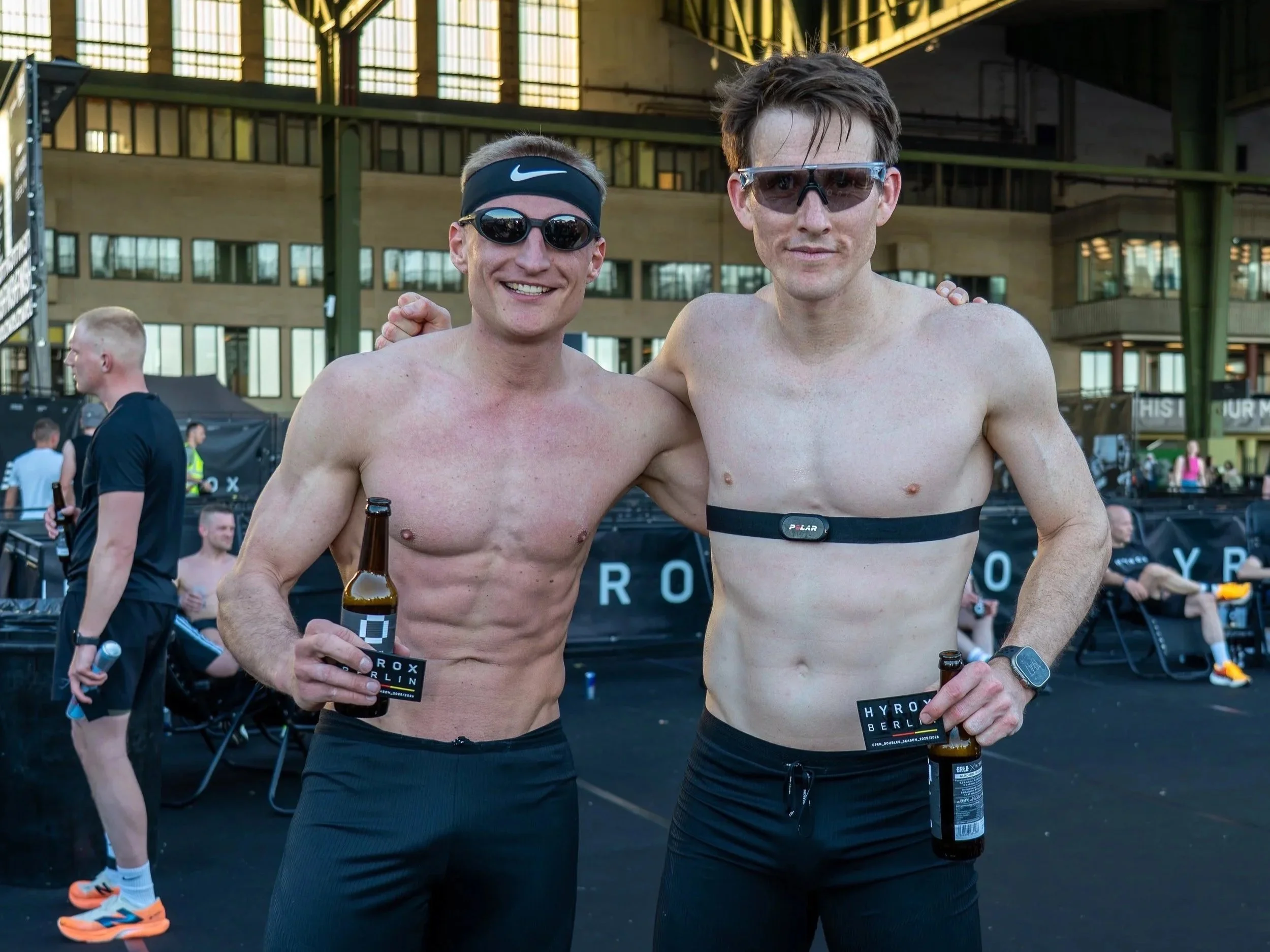 Two shirtless male athletes wearing sunglasses and black shorts, holding beer bottles, smiling at the camera inside a sports venue with an industrial ceiling and large windows, surrounded by other athletes and spectators.