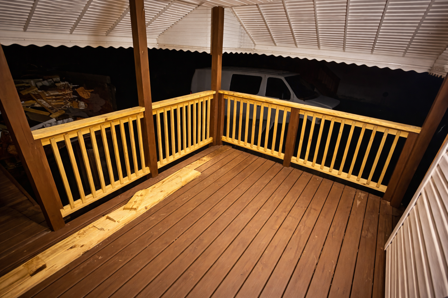 A newly built wooden balcony with a railing, illuminated by warm lighting, overlooking a dark yard or driveway with a parked van and some storage items.