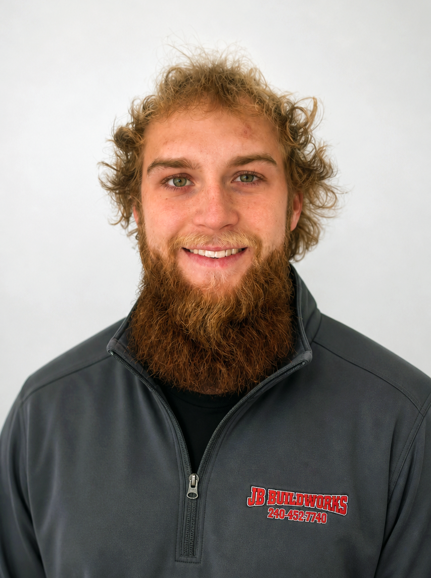 A young man with curly blonde hair and a full beard smiling, wearing a gray jacket with a JB Buildworks logo.