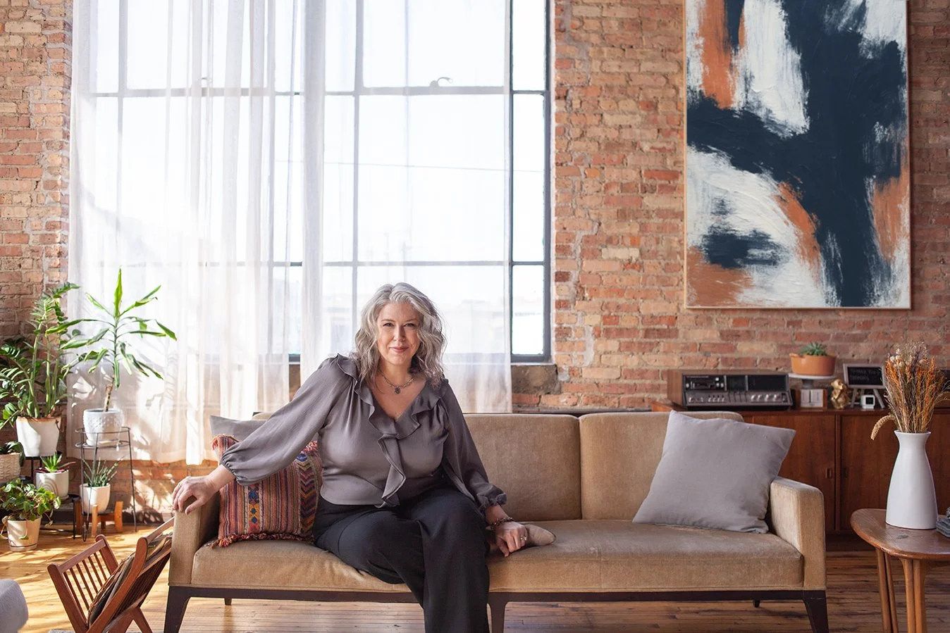 A middle-aged woman with gray hair sitting on a beige sofa in a modern, industrial-style living room with large windows, brick walls, and contemporary art on the wall, surrounded by indoor plants.