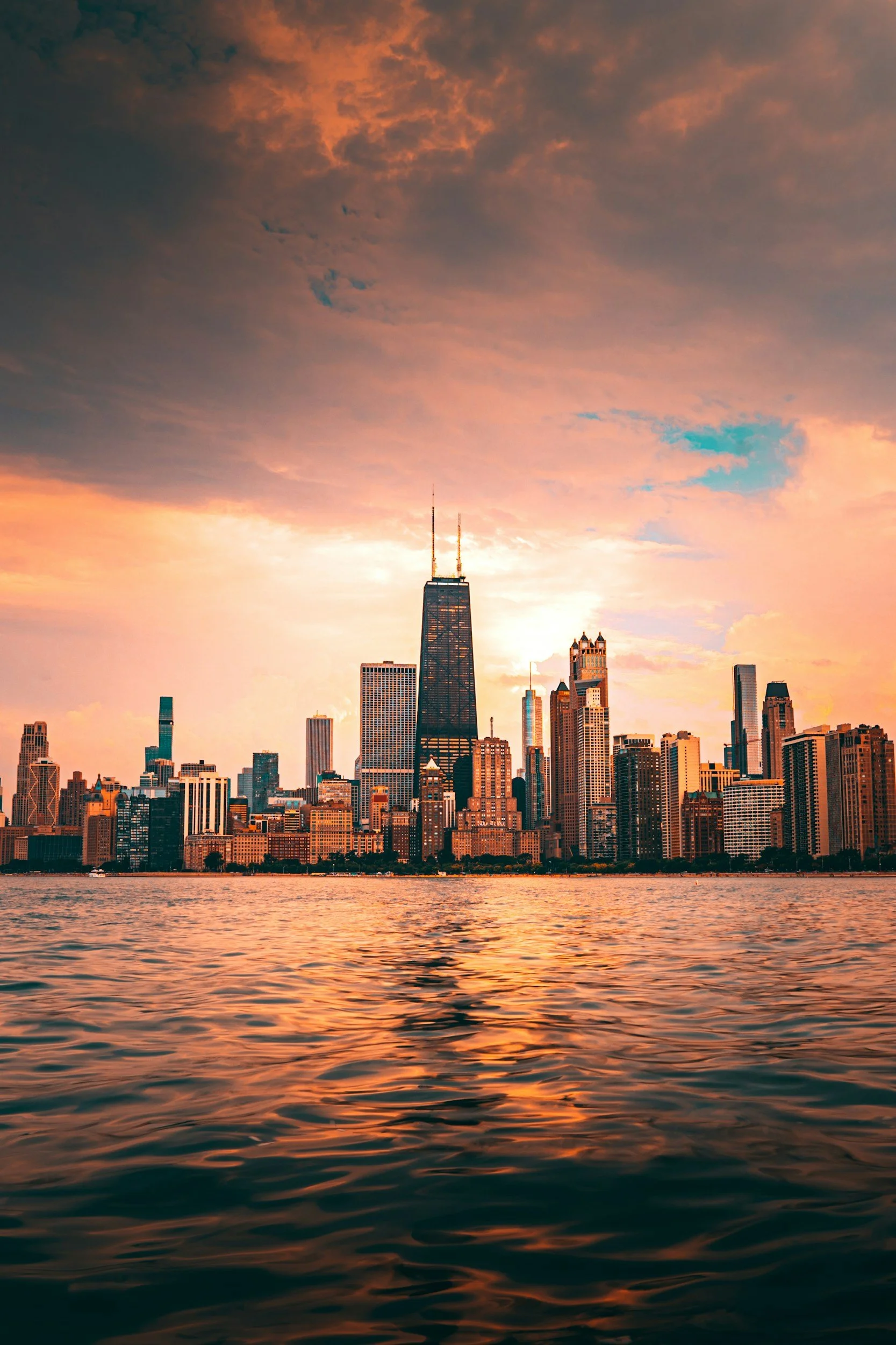 A city skyline during sunset with tall buildings, including the Willis Tower, reflected in a body of water in the foreground, and a colorful sky with clouds.