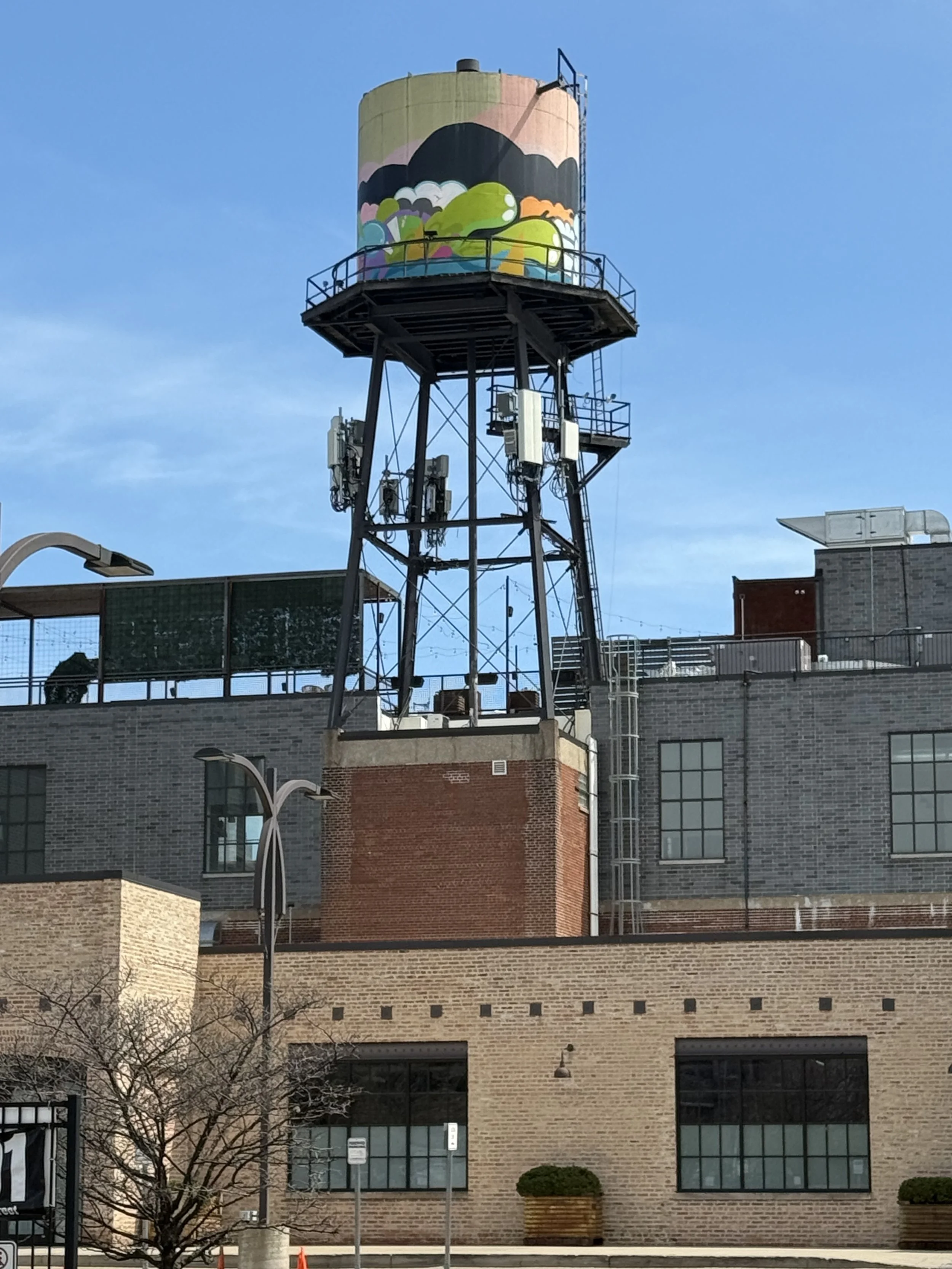 A West Loop water tower with a colorful mural featuring clouds, green hills, and trees, situated above brick and concrete buildings with windows and utility installations.
