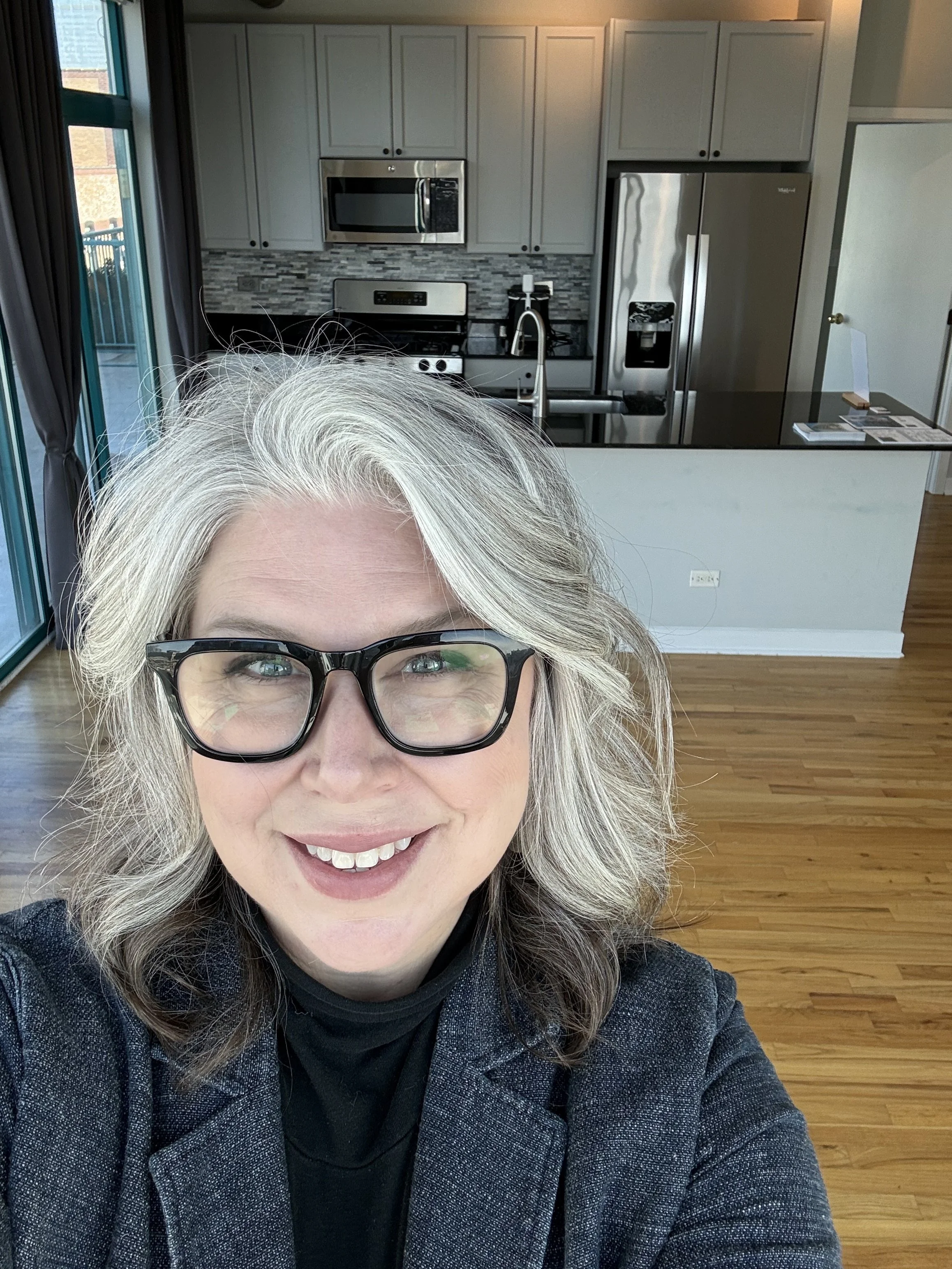 A woman with gray hair wearing black glasses and a dark blazer, smiling for a selfie in a modern kitchen with stainless steel appliances and light wood flooring.