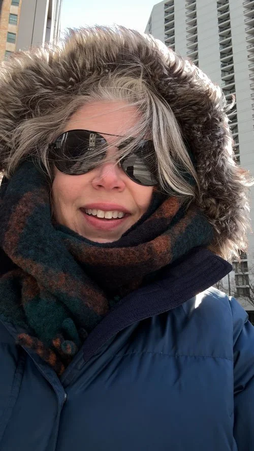 Woman wearing sunglasses and a winter coat with a fur-lined hood, smiling outdoors against city buildings.