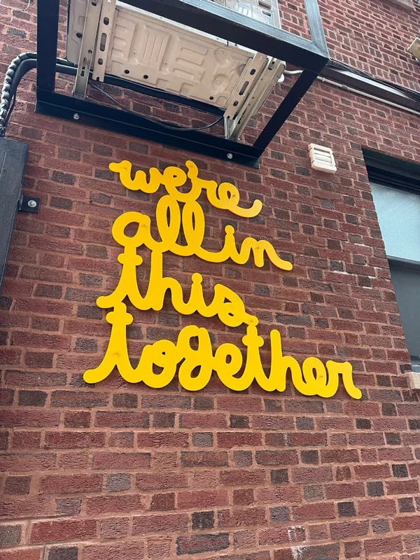 Yellow cursive sign on a brick wall reads "we all in this together".