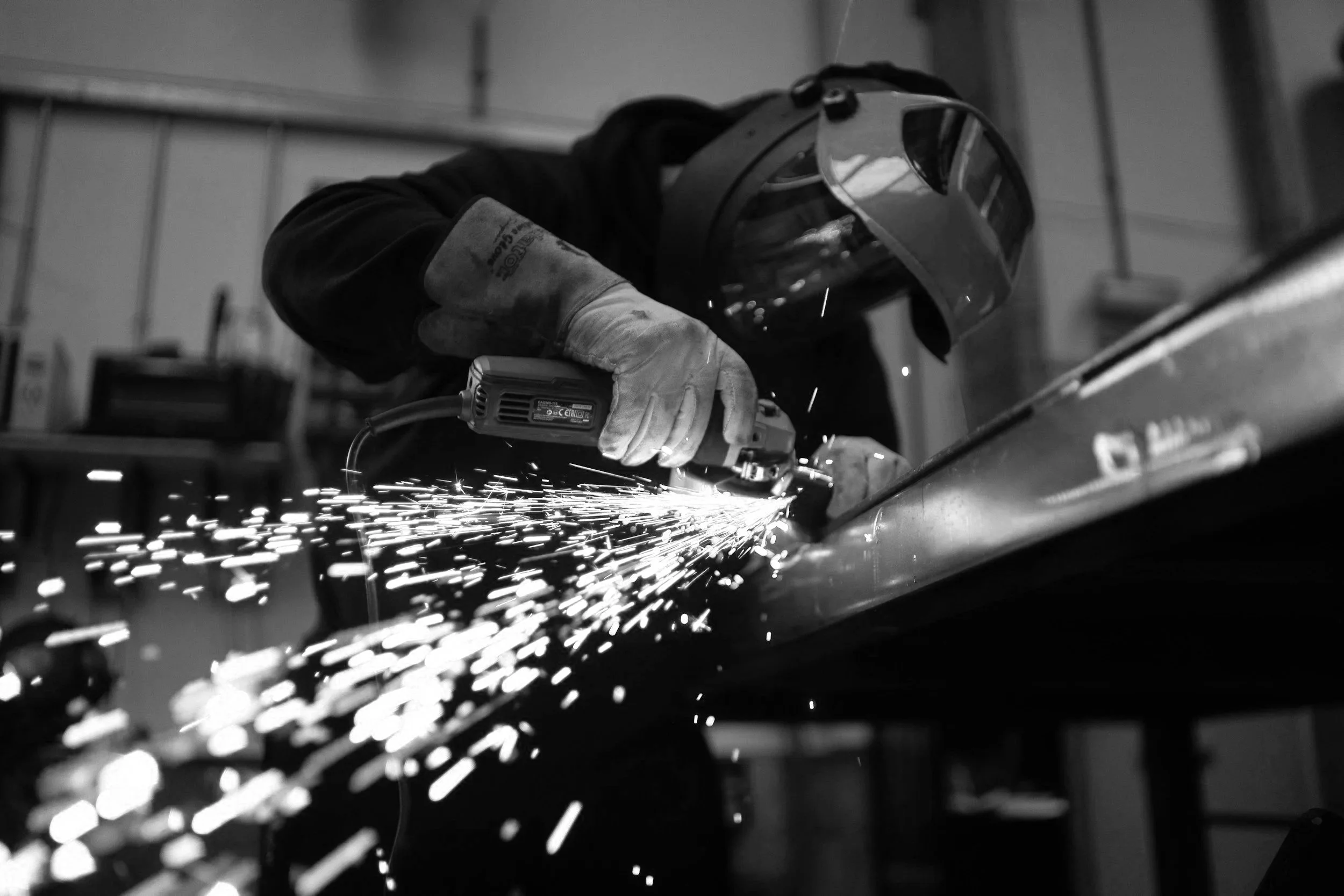 A person wearing a welding helmet and gloves, using an angle grinder to cut or grind a metal piece, creating sparks in a workshop.