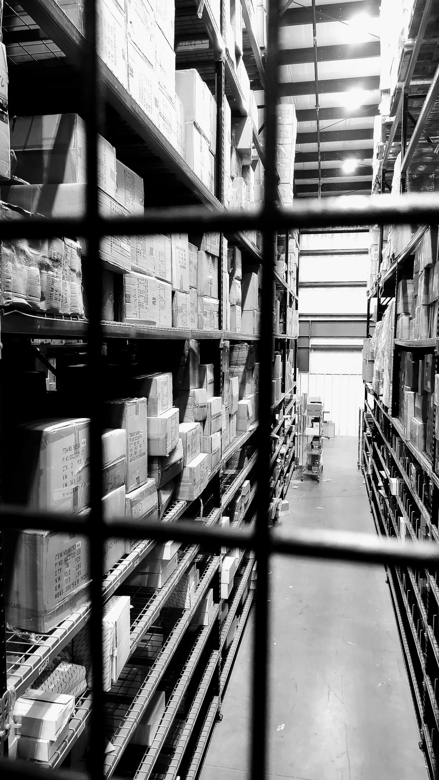 View through a wire fence into a warehouse aisle filled with shelves stocked with boxes and packages