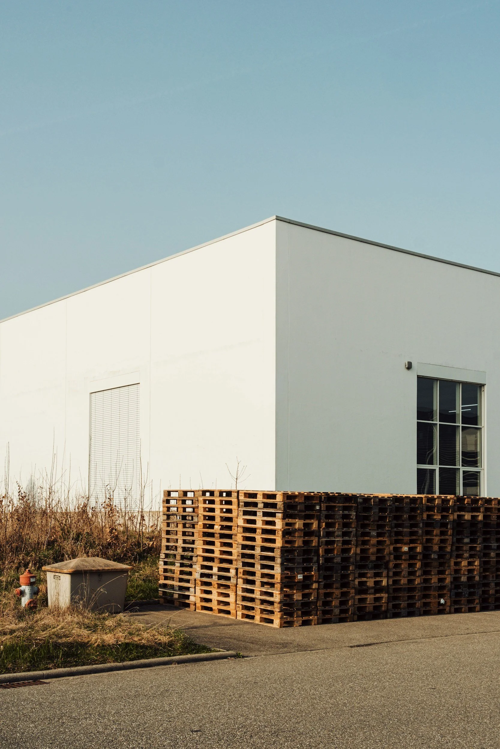 A white modern building with a large window and a window with closed blinds, beside a pile of stacked wooden pallets on the sidewalk, and a fire hydrant to the left.