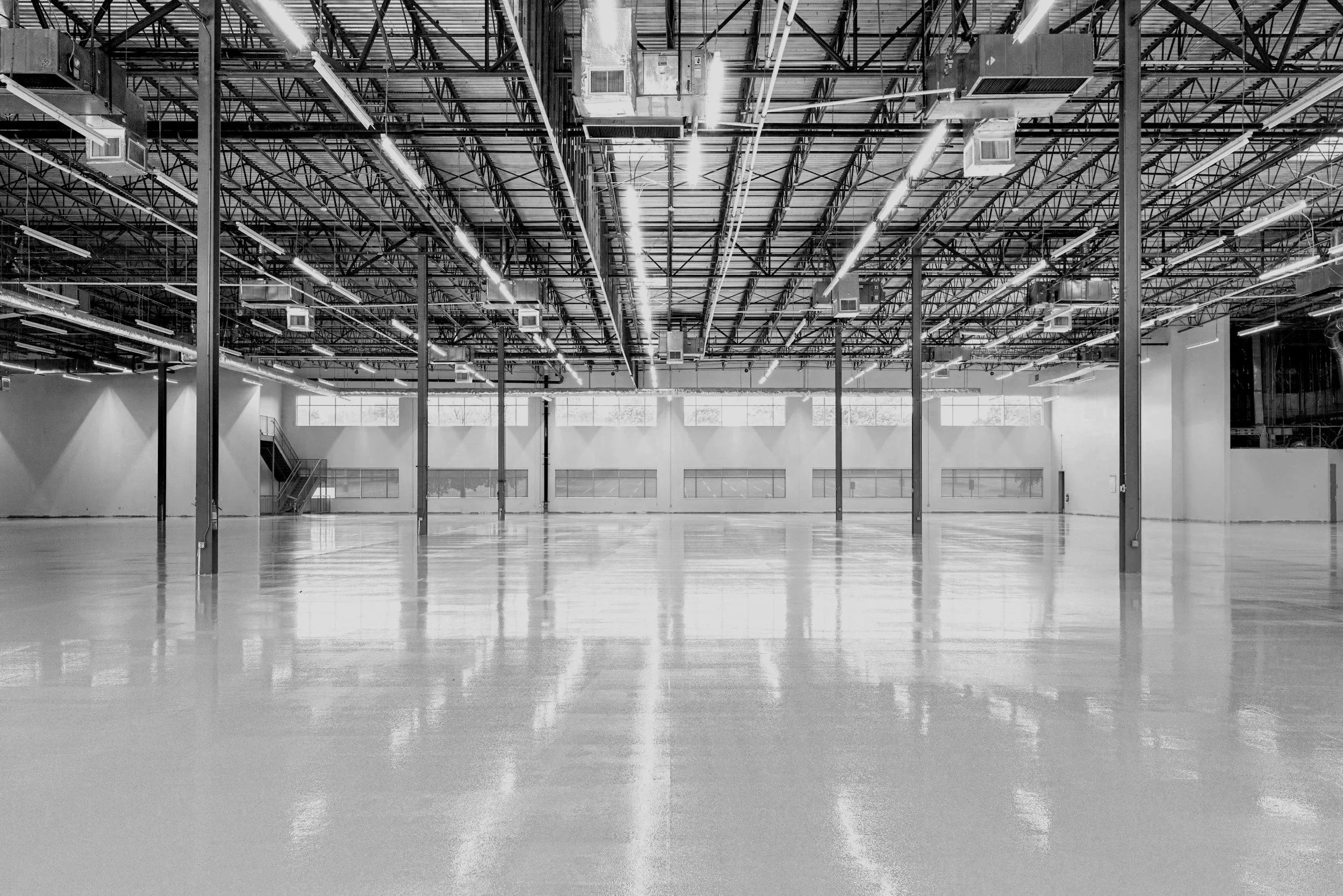 Empty industrial or warehouse space with polished floor, exposed ceiling with structural beams, pipes, and lighting fixtures, and large windows allowing in natural light.