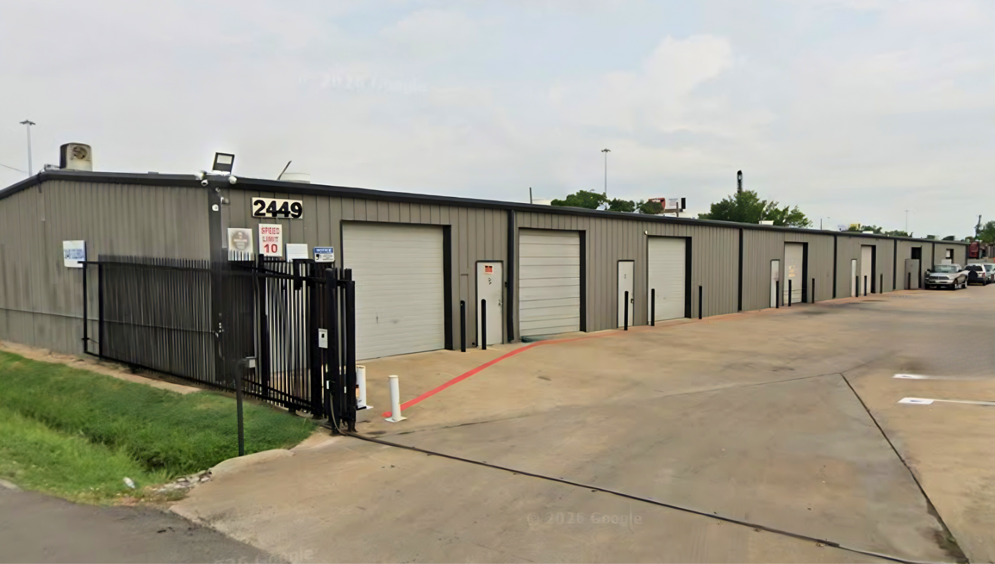 A row of storage units with roll-up doors in a storage facility, with cars parked on the right side and a chain-link fence gate at the front.