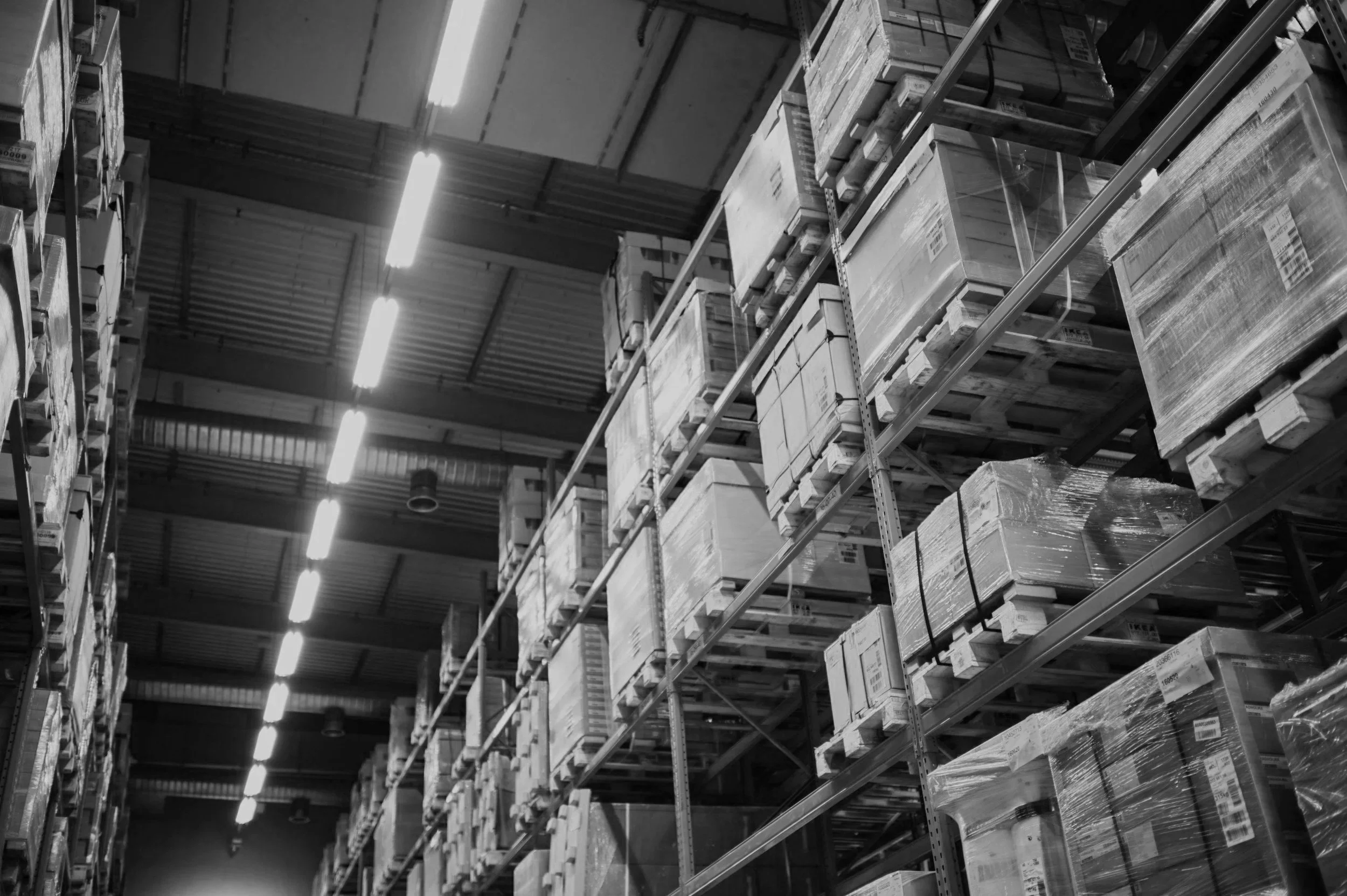 Black and white photo of warehouse shelves loaded with boxes and pallets, illuminated by overhead fluorescent lights.
