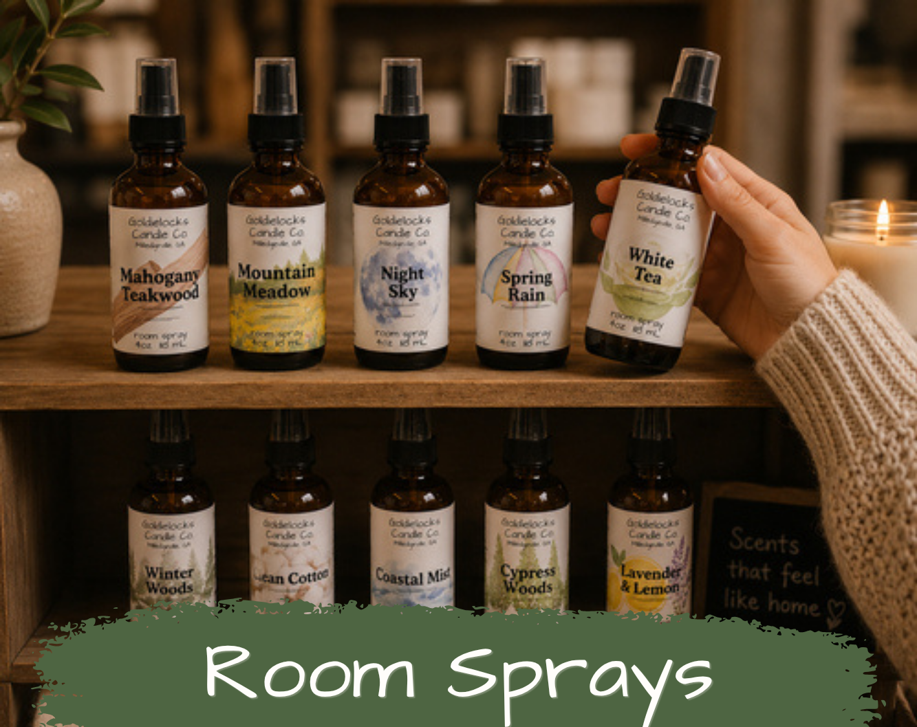 A hand holding a spray bottle labeled 'White Tea' among five other room spray bottles with labels: Mahogany Teakwood, Mountain Meadow, Night Sky, Spring Rain, and White Tea, on a wooden shelf with a lit candle and a plant in the background.