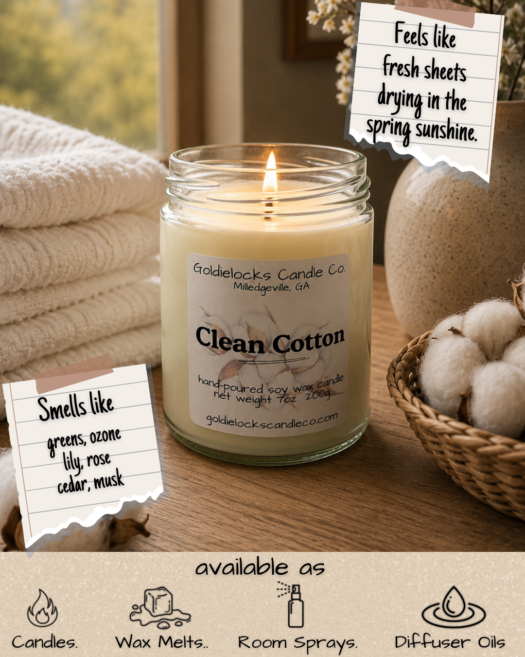 A lit scented candle labeled 'Clean Cotton' by Goldielocks Candle Co., sitting on a wooden surface with folded towels, cotton balls, and a small basket. Handwritten notes mention it smells like greens, ozone, lily, rose, cedar, and musk and feels like fresh sheets drying in spring sunshine.
