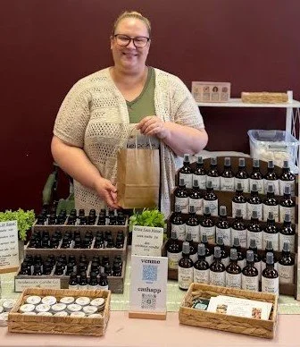 Woman smiling at a booth displaying essential oil bottles, informational cards, and room sprays on a table with a burgundy wall background.